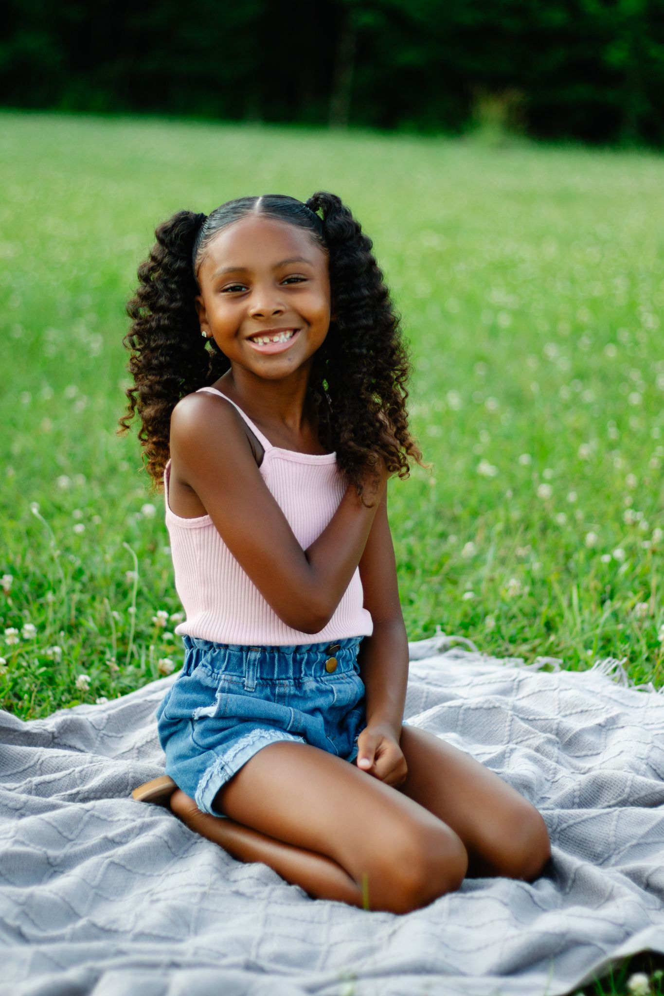 Child in denim overalls sitting on blanket in grassy field on sunny day.