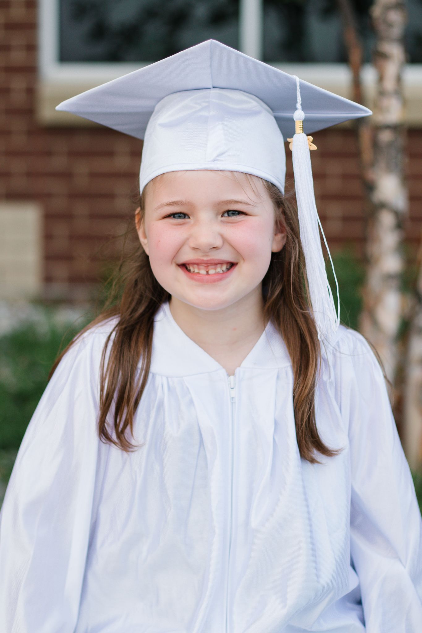 Graduate in white cap and gown poses outside a brick building on graduation day.