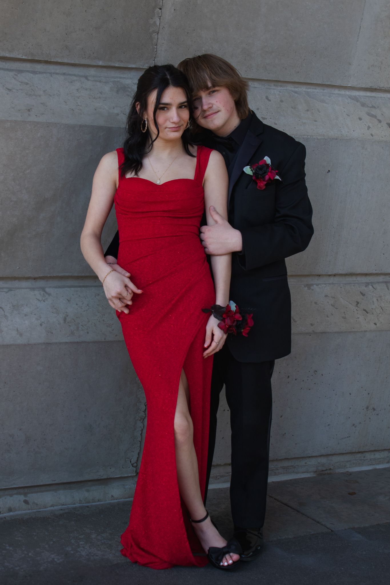 A couple poses together in formal attire with one person wearing a vibrant red gown against a stone wall.