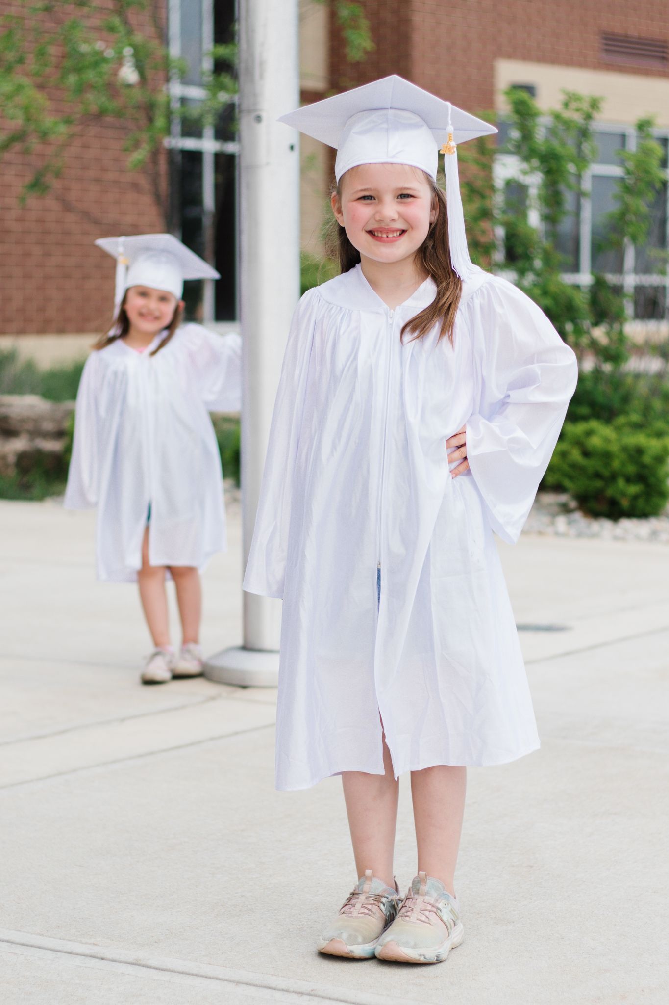 Graduate in white cap and gown stands on sidewalk near brick building.
