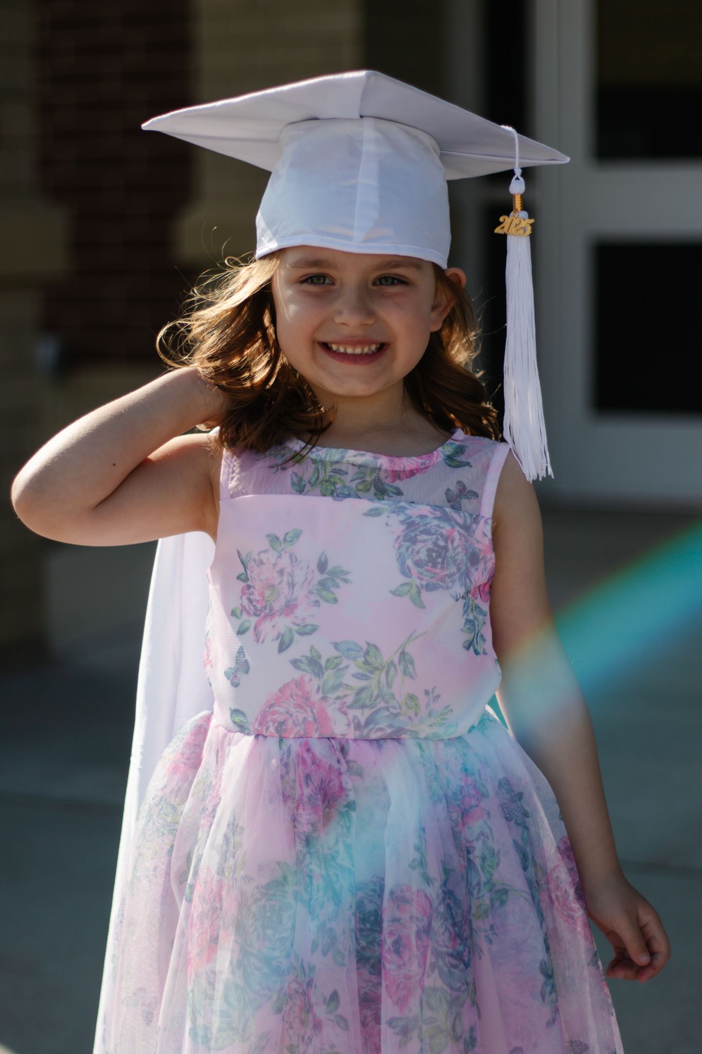 A lovely graduate in a pink and blue floral dress and white cap smiles outdoors.