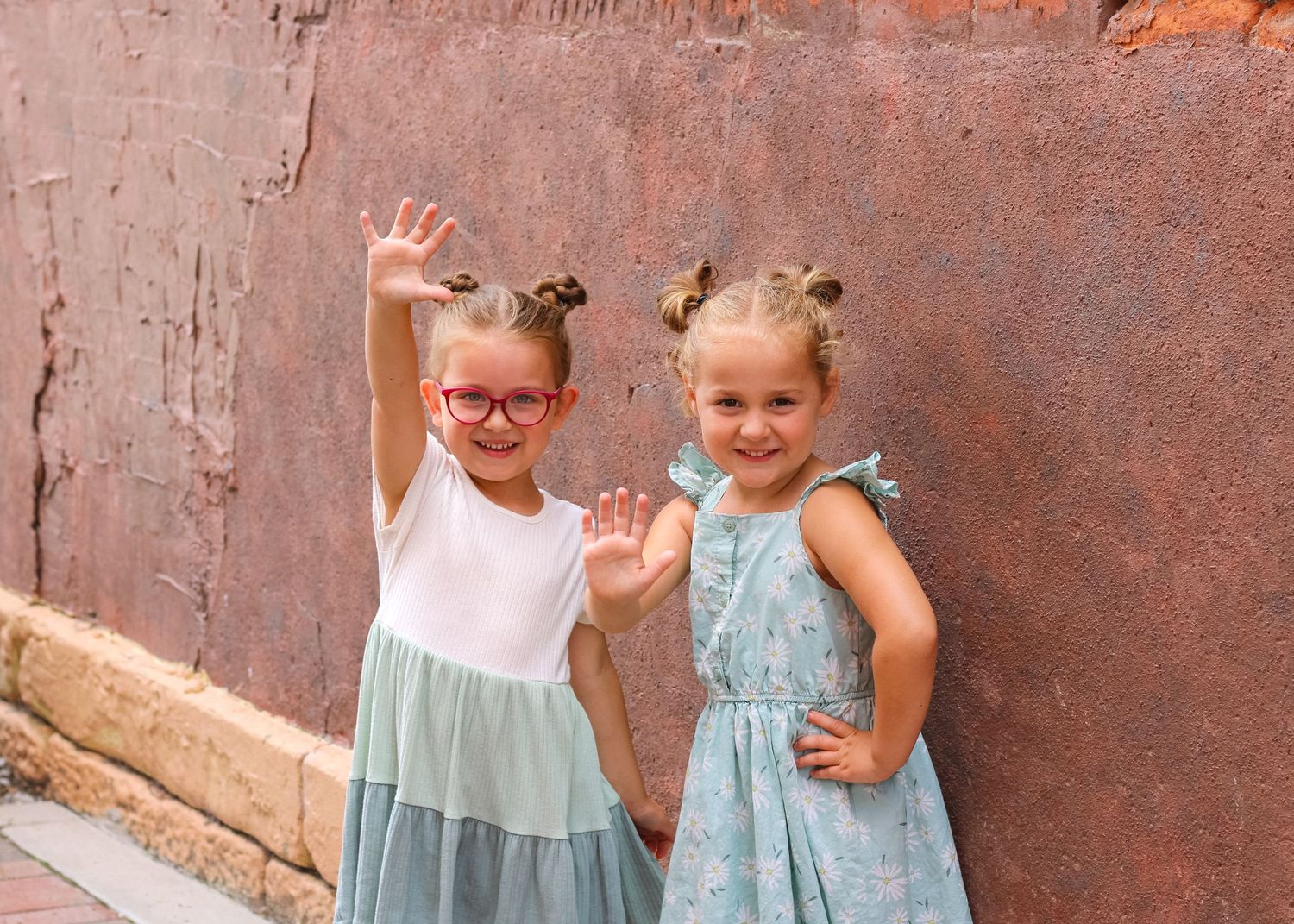 Children posing against rustic orange wall wearing coordinated blue and pink dresses.