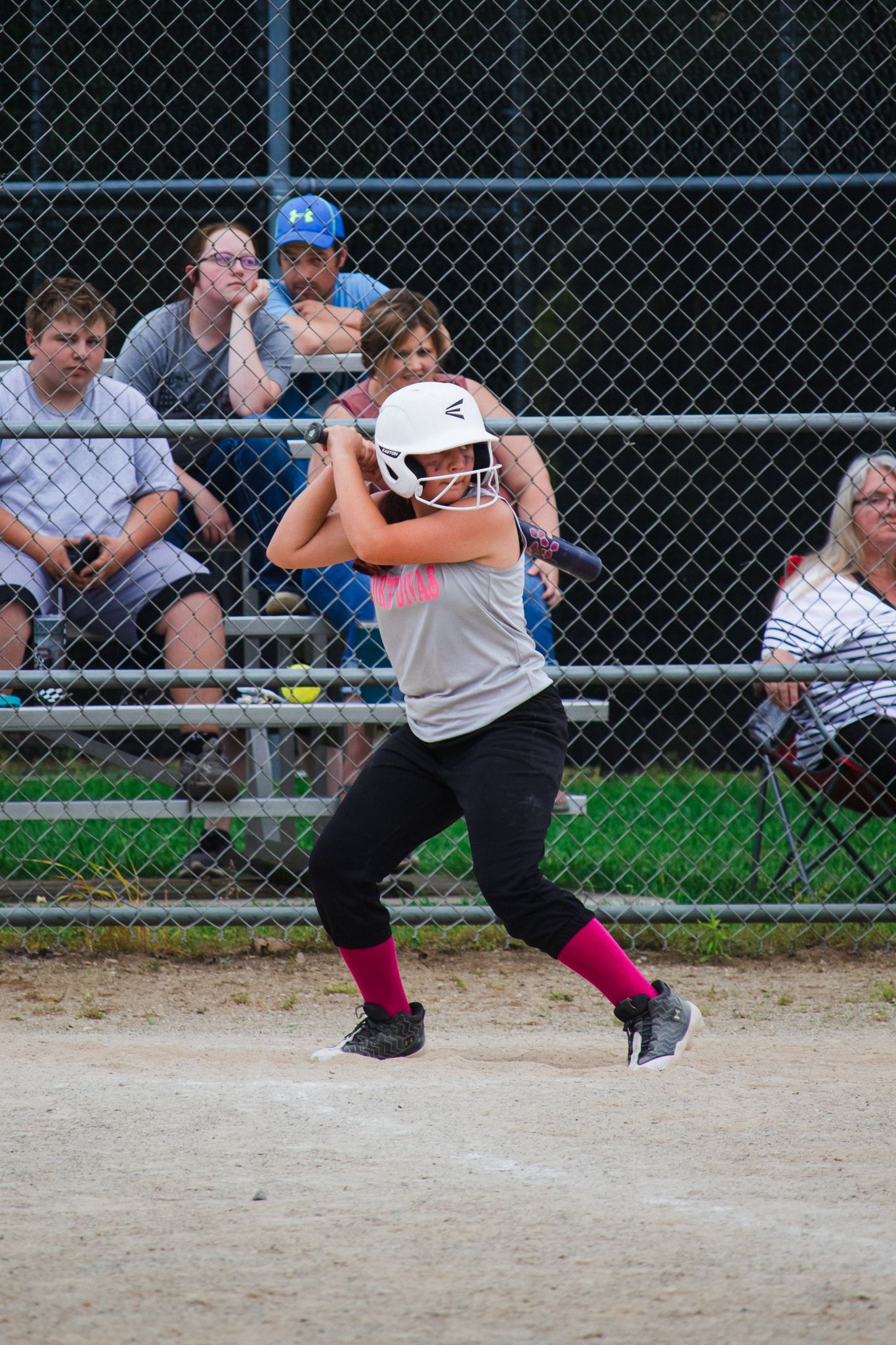 Wide action shot of softball batter mid-swing with spectators watching through fence.
