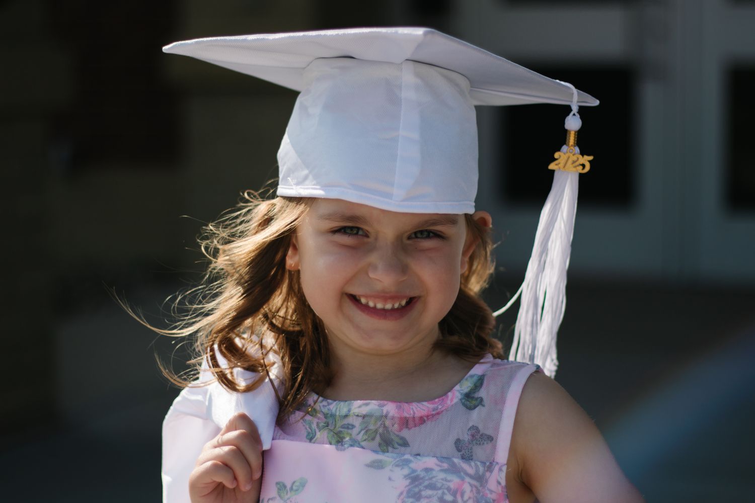 Graduate in white cap and gown smiling outdoors in summer sunlight.