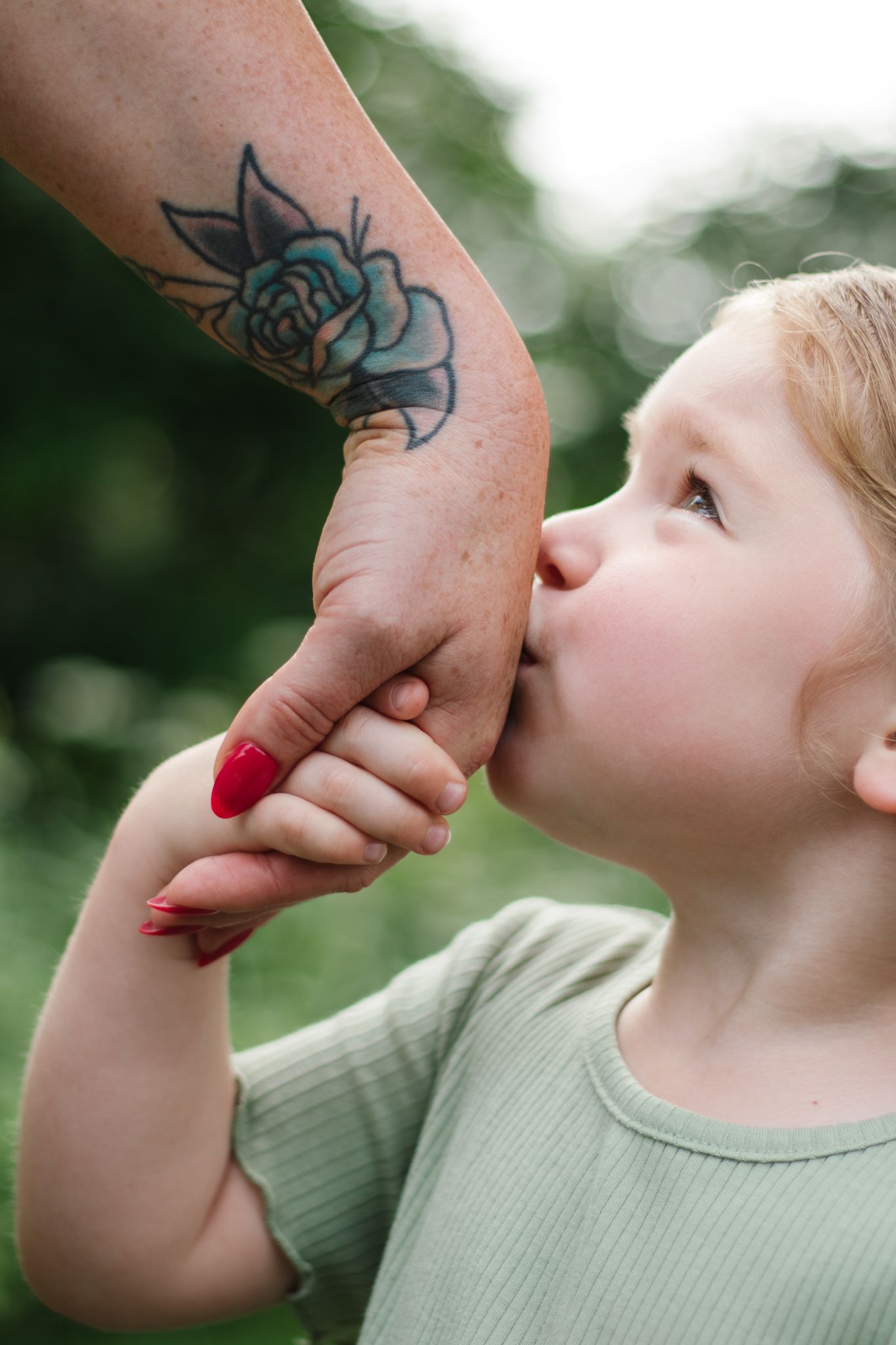 A small child in a mint green shirt kisses a tattooed hand against a blurred background.