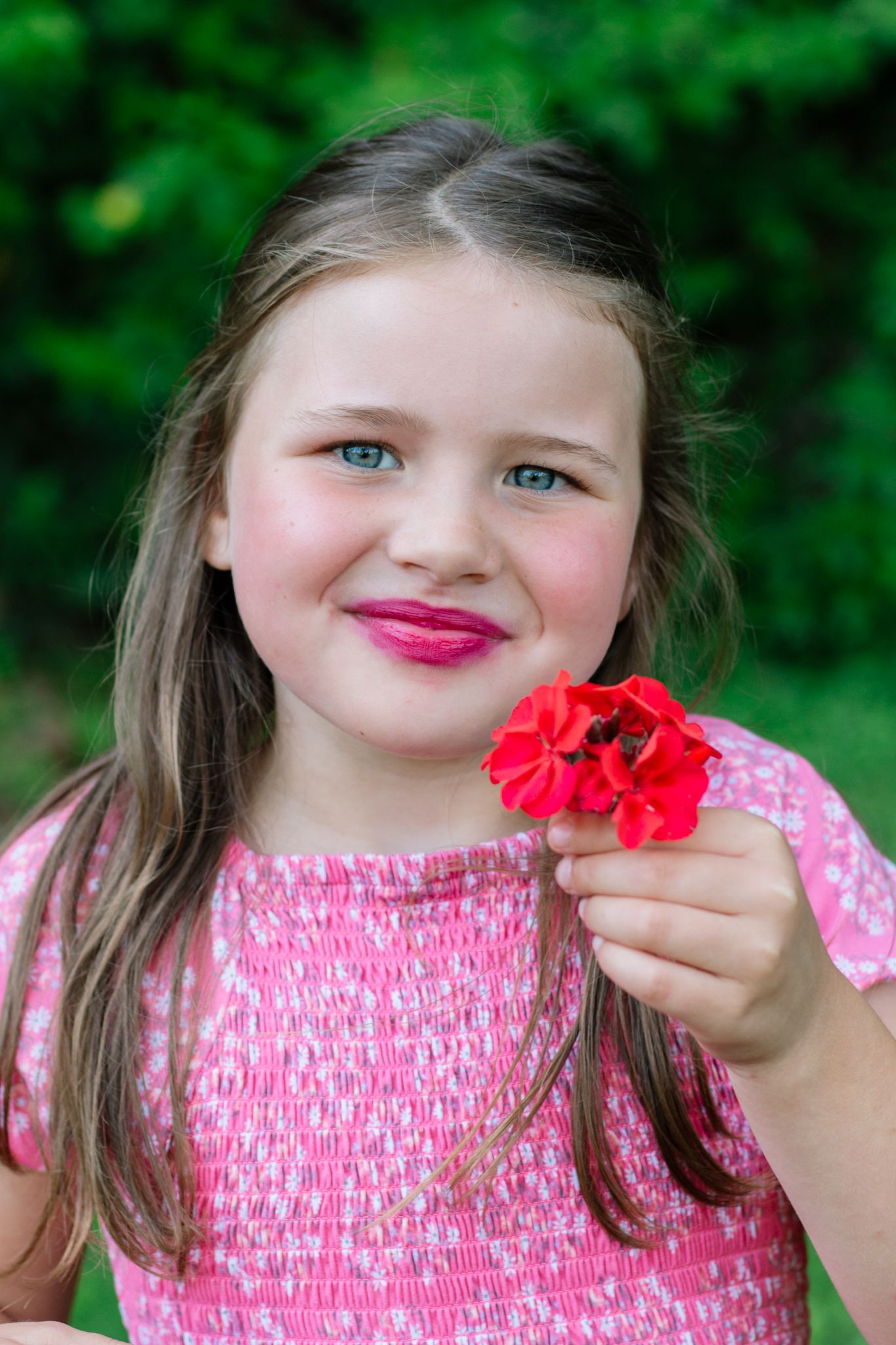 Young person in pink smocked top holding red flower against green background.