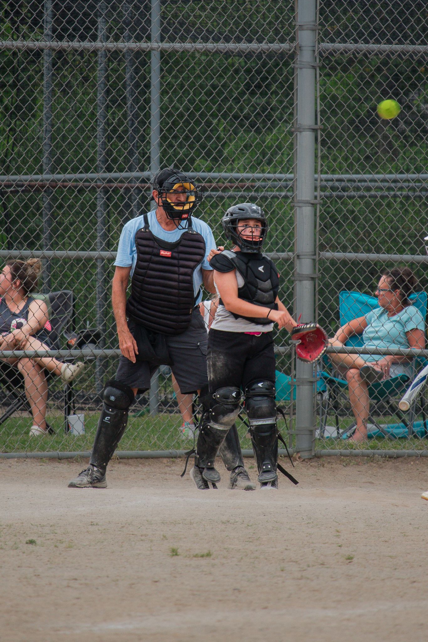 Wide shot of softball game with catcher, umpire and spectators behind chain link fence.