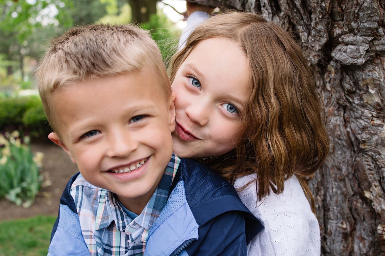 Two children sharing sweet candid moments while sitting near large tree.
