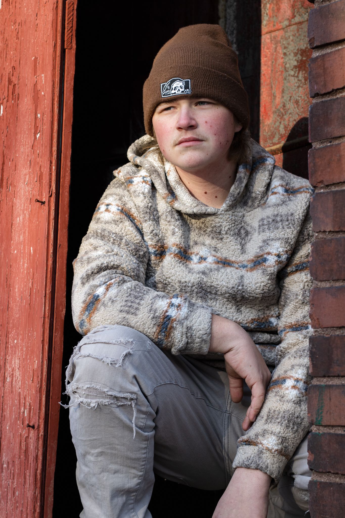 Figure wearing beanie and patterned sweater poses against red painted brick wall.