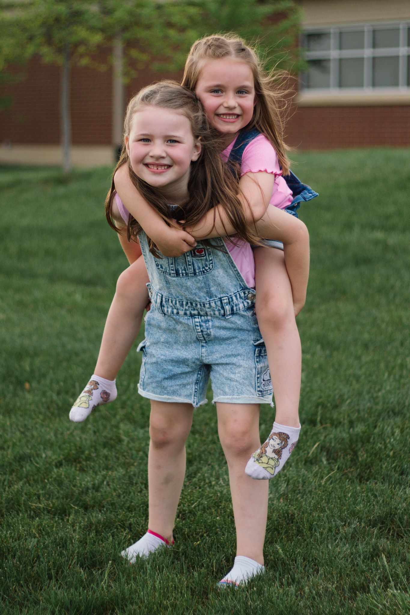 Child playing and bending over on grass wearing denim overalls and pink shirt.