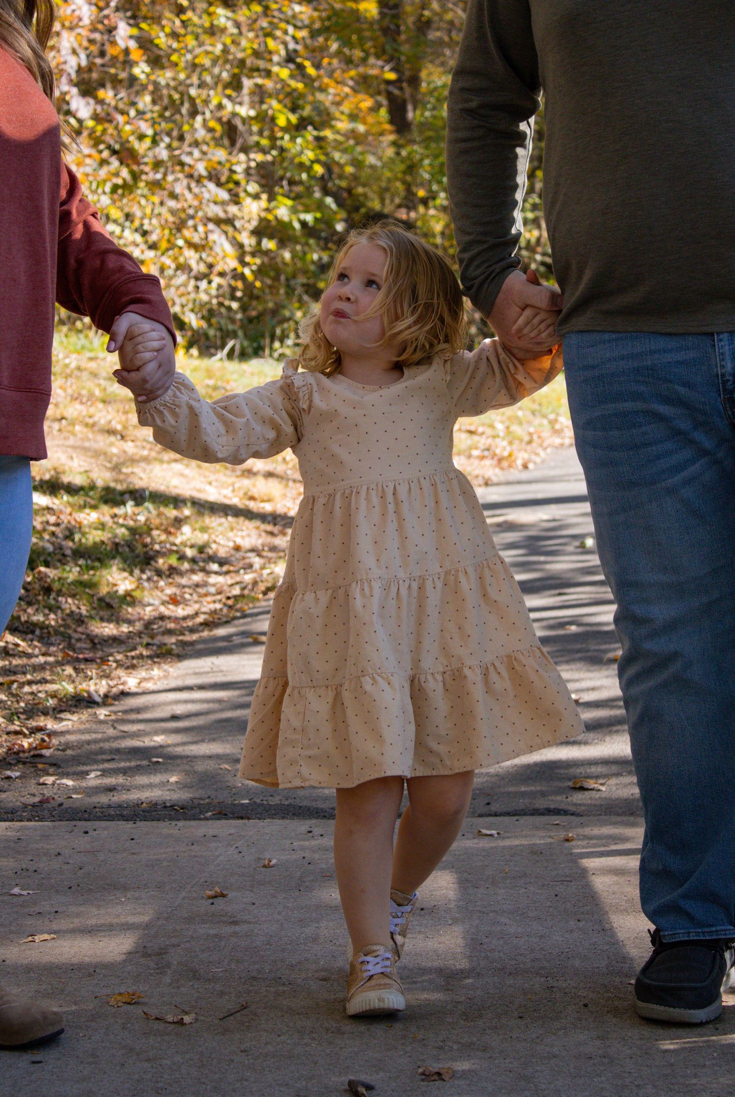 Young child in a cream-colored dress twirls playfully on a path surrounded by autumn foliage.