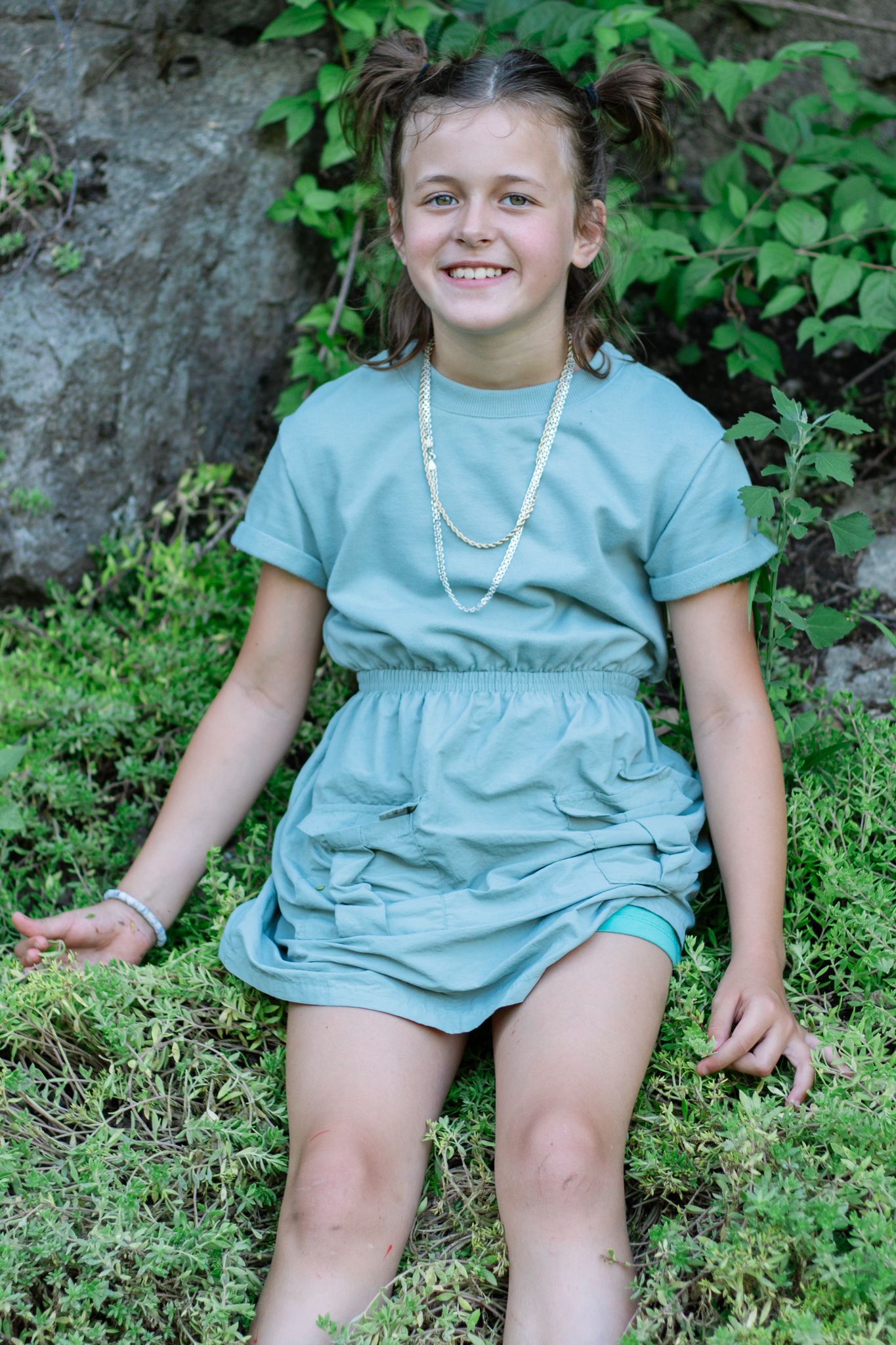 A figure in mint green clothing sits casually on grass and rocks in a garden setting.