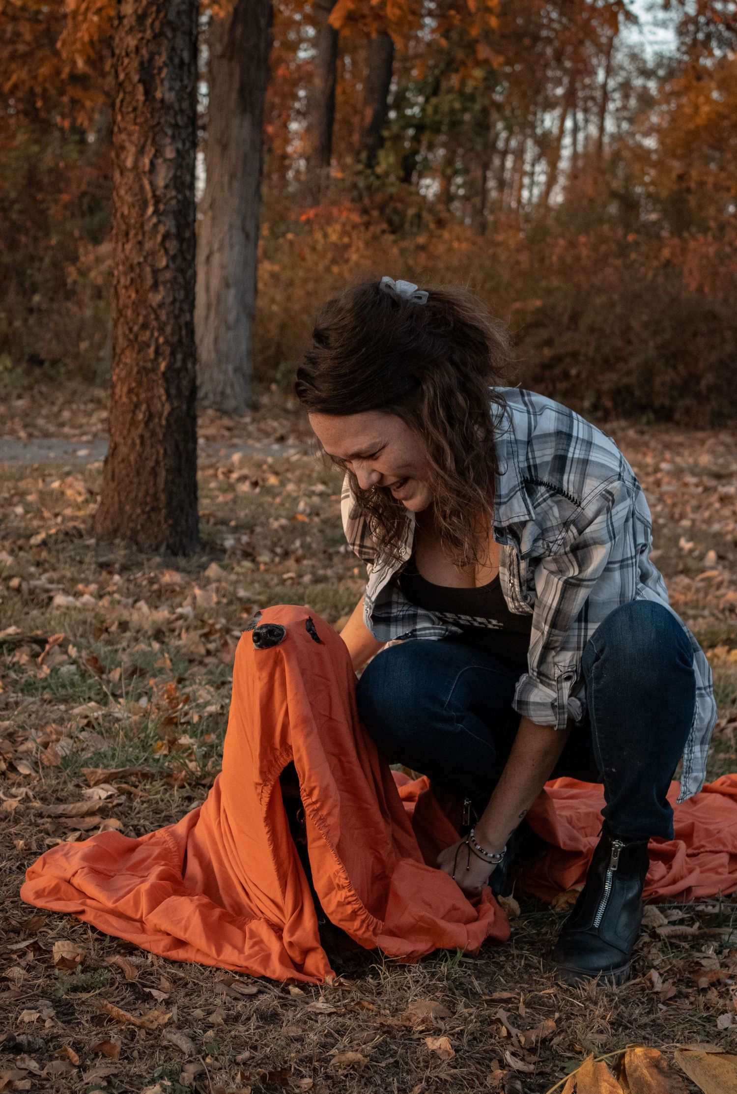 Person in plaid shirt interacting with orange fabric on fallen leaves in wooded area.