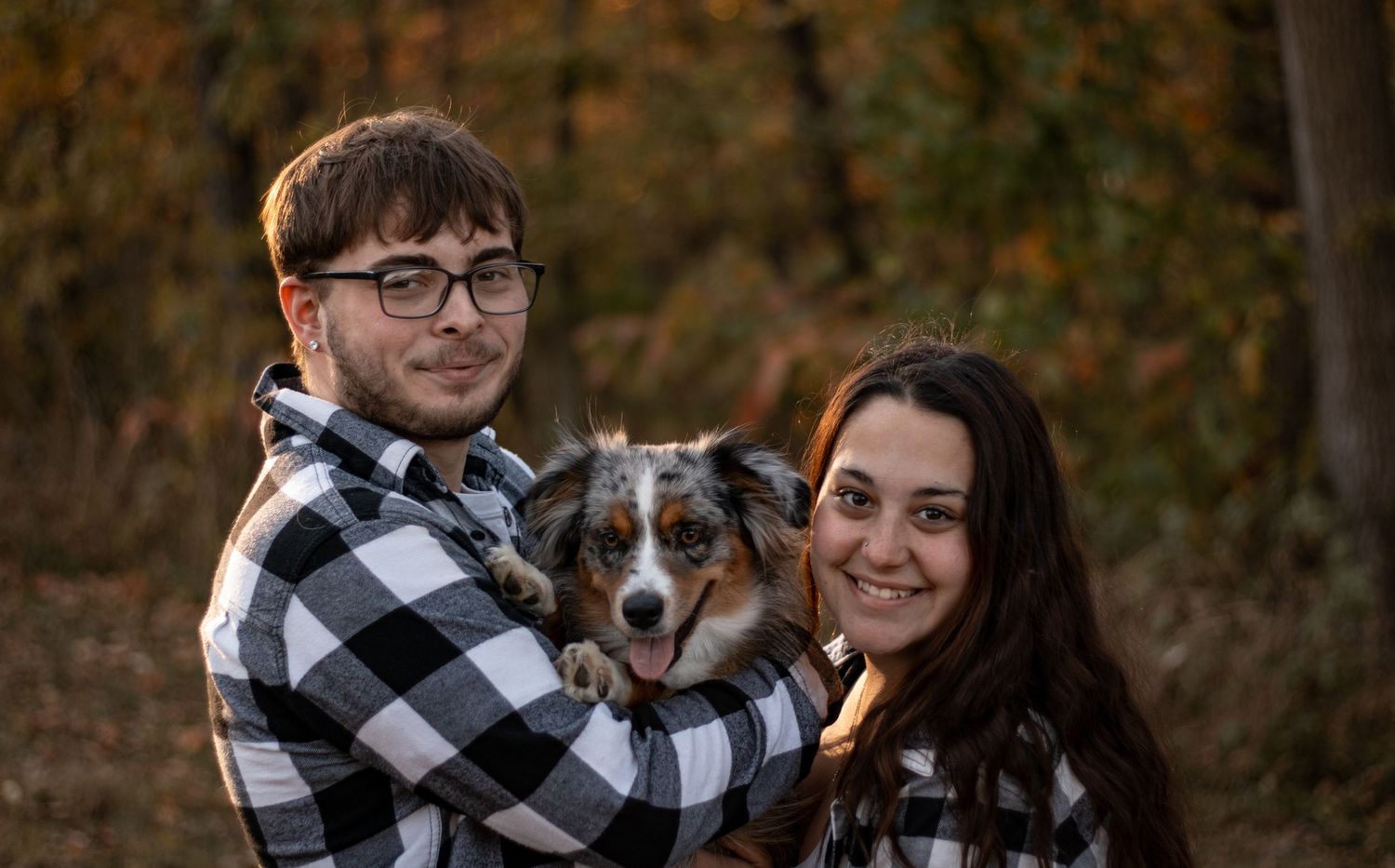 Couple poses with their Australian shepherd dog against a fall foliage background.