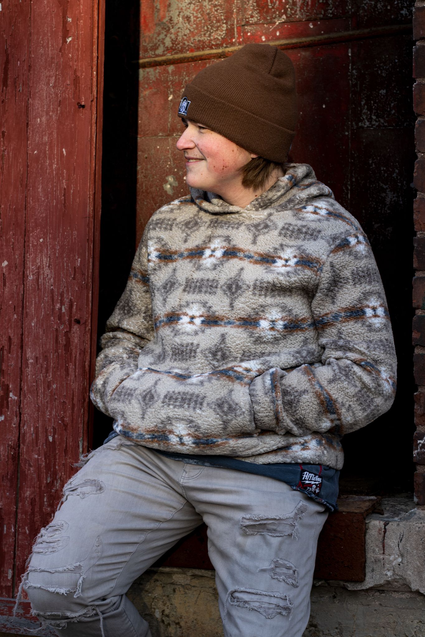 Person in patterned sweater and gray pants sits casually by red painted wall.