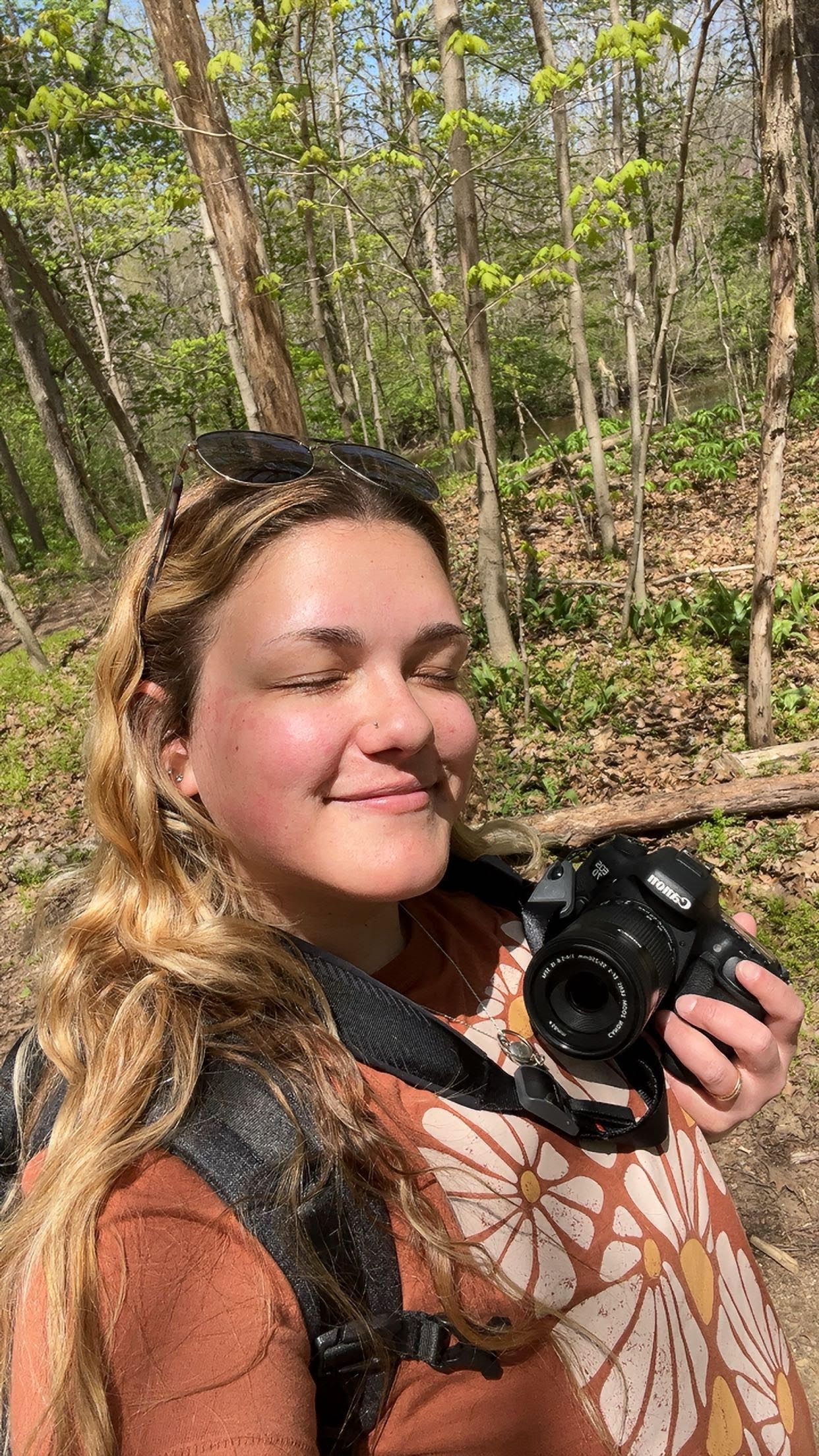 Photographer with wavy hair holding camera outdoors among trees in natural lighting.