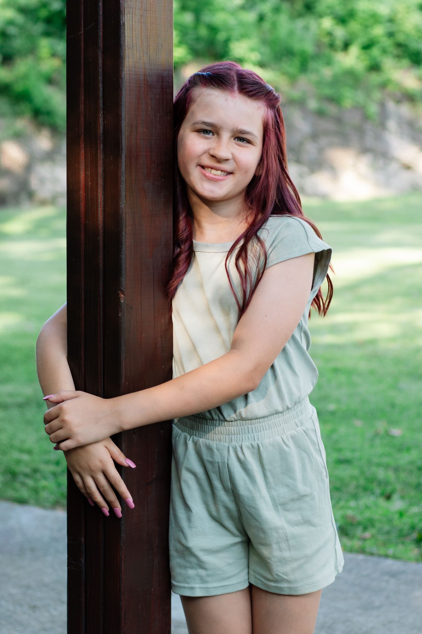 A person in a sage green romper poses playfully against a wooden post in an outdoor setting.