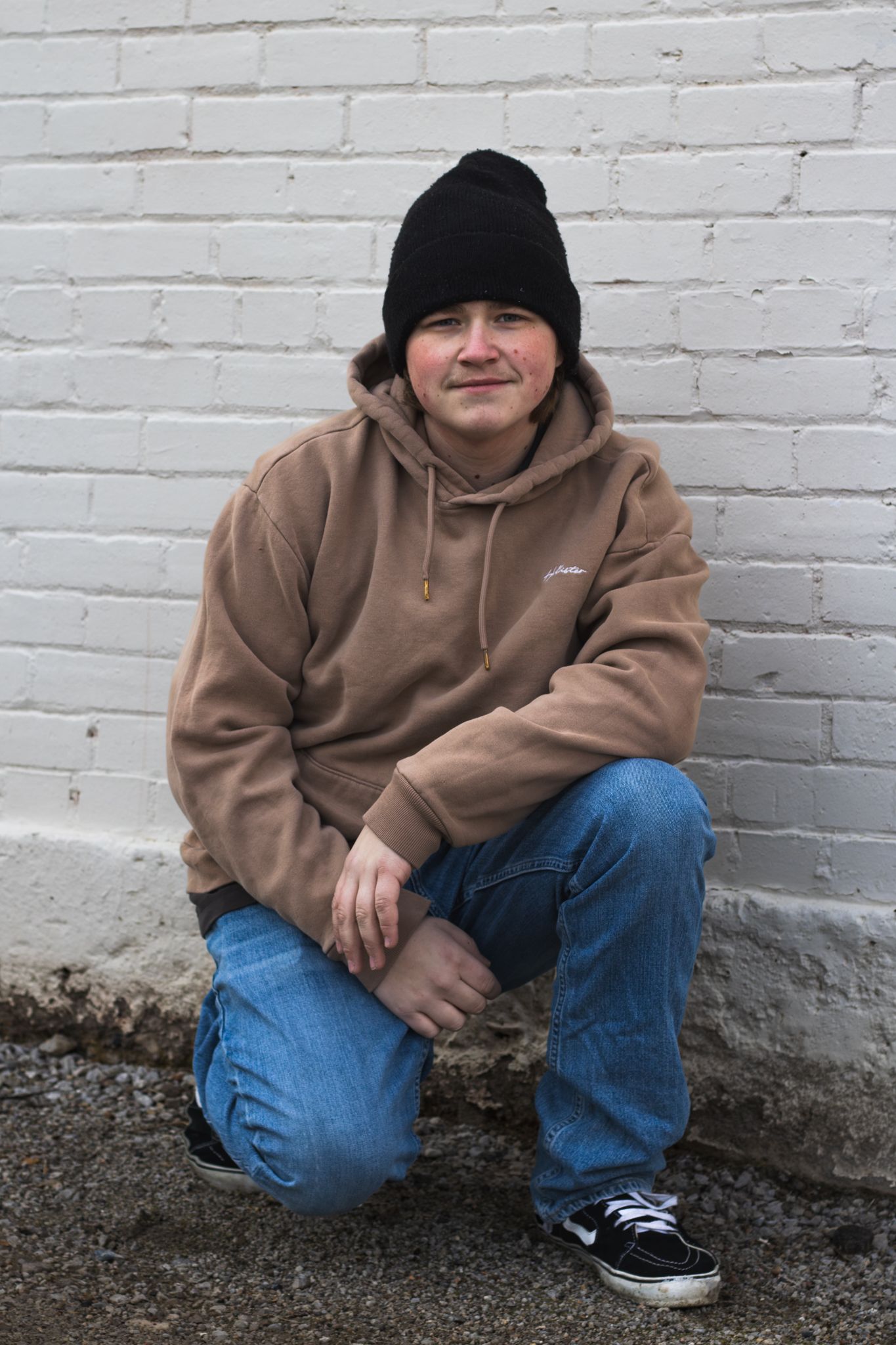 Person in brown hoodie and jeans sits against white brick wall.