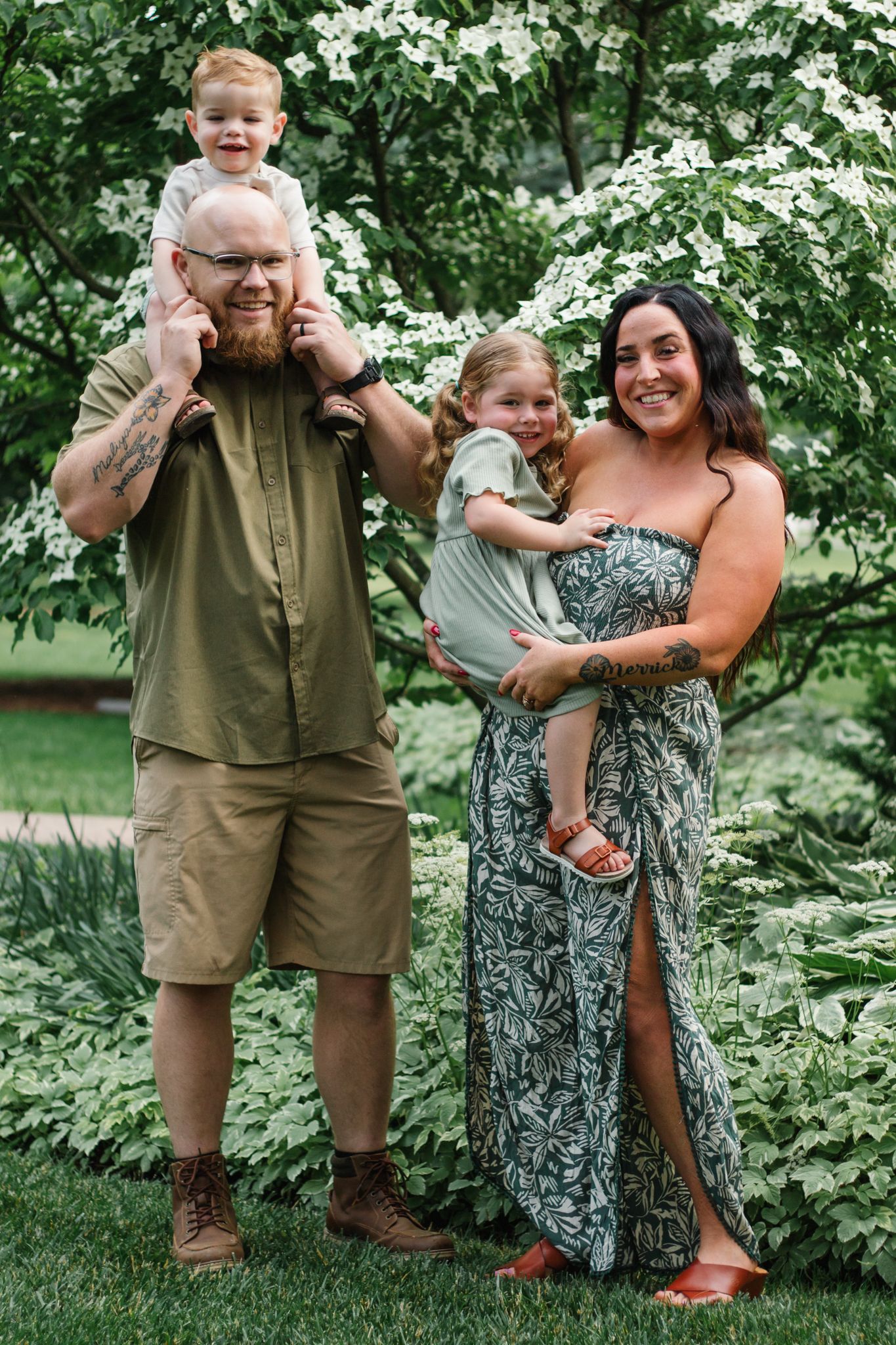 Family wearing coordinated olive and neutral outfits poses playfully in a green outdoor garden setting.
