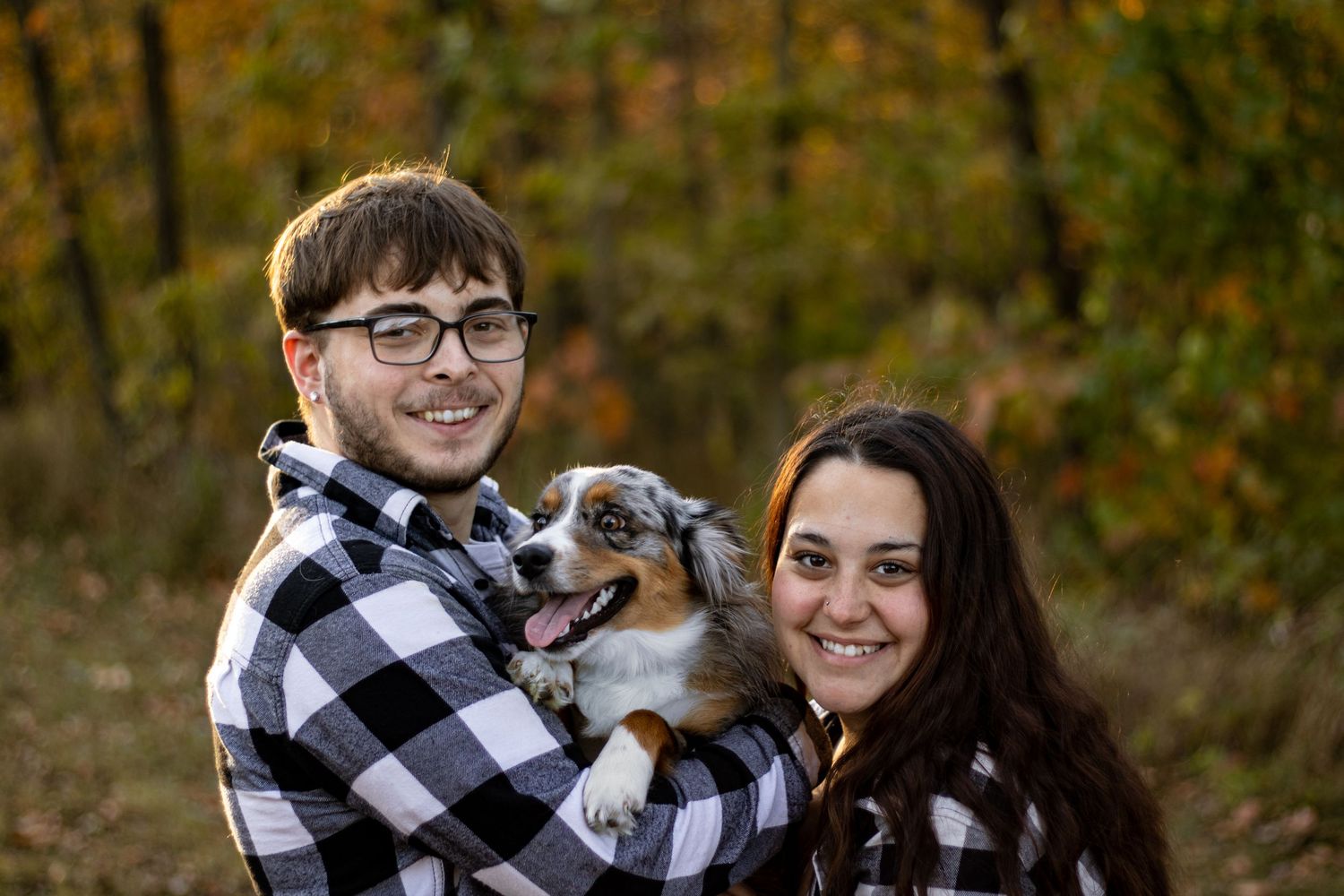 Happy couple in buffalo plaid holds their Australian shepherd puppy in an autumn setting.