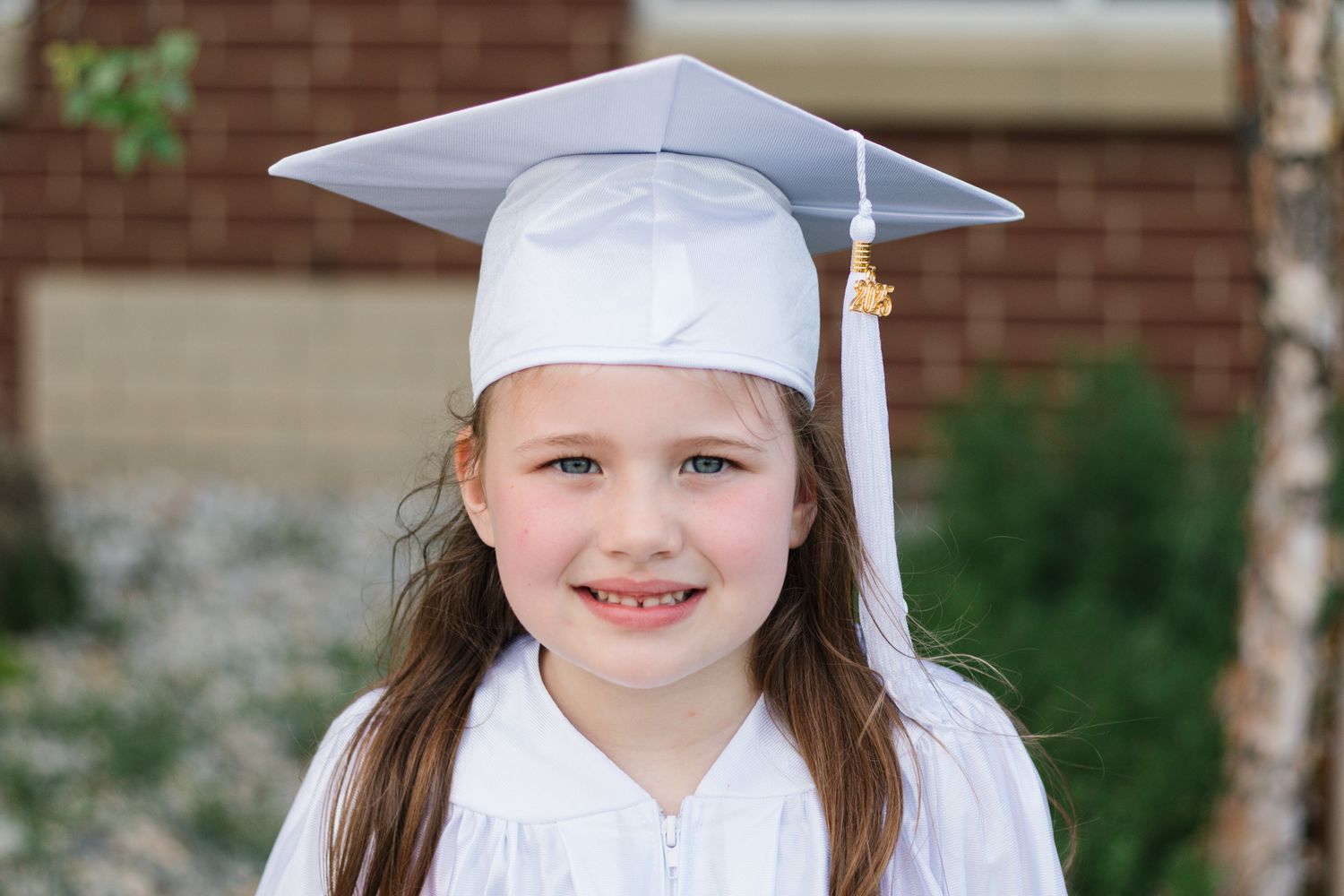 Graduate in traditional white cap and gown poses against brick wall background.