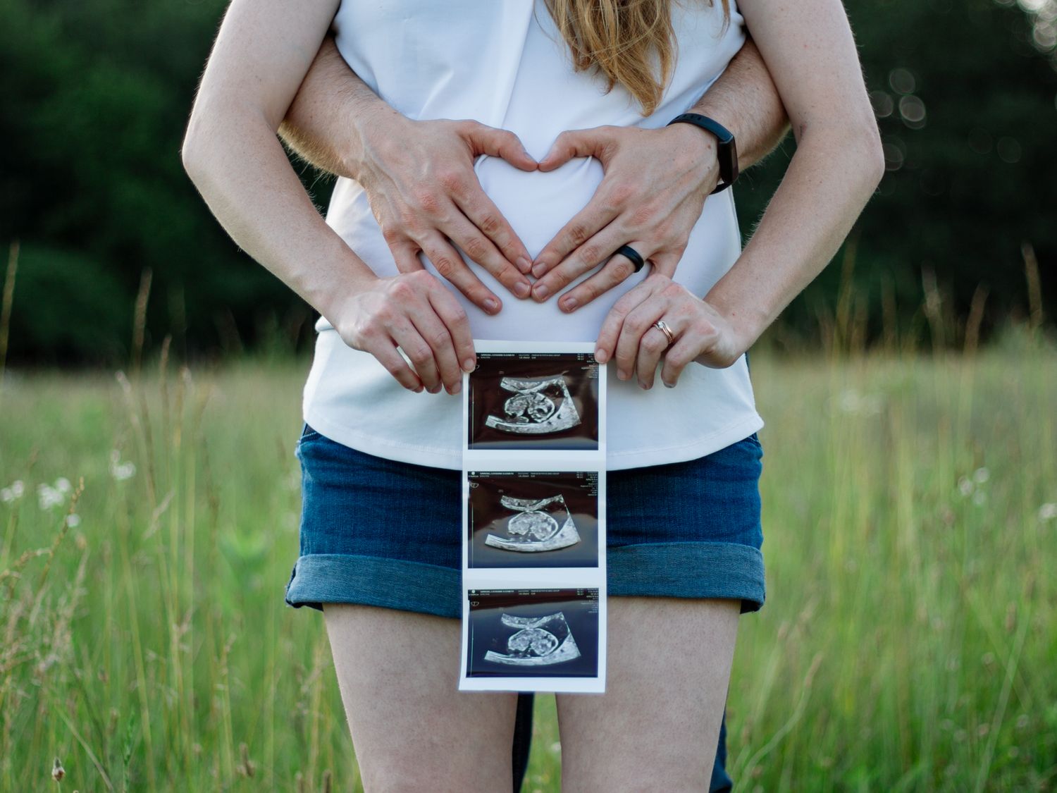 Series of hands making heart shapes around ultrasound photos in grassy field.