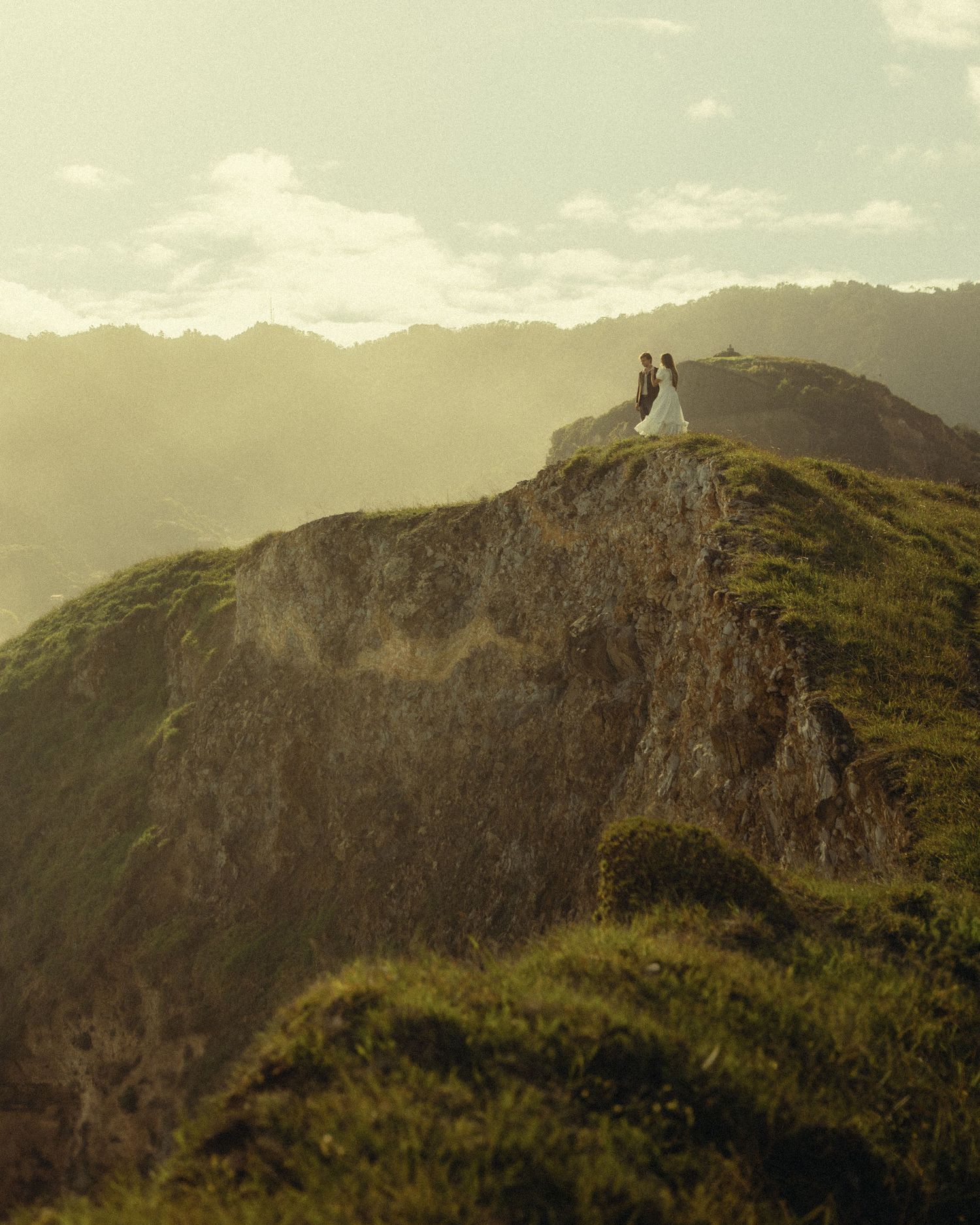 A couple in wedding attire stands atop a dramatic mountain cliff overlooking lush green valleys and peaks at sunset.
