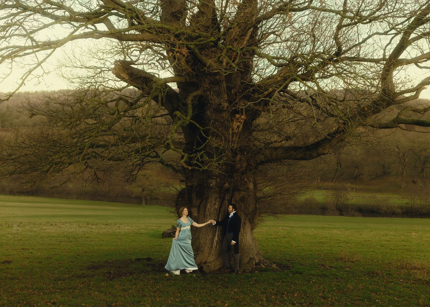 A couple in period attire stands beneath a large bare tree in a lush green field during overcast weather.