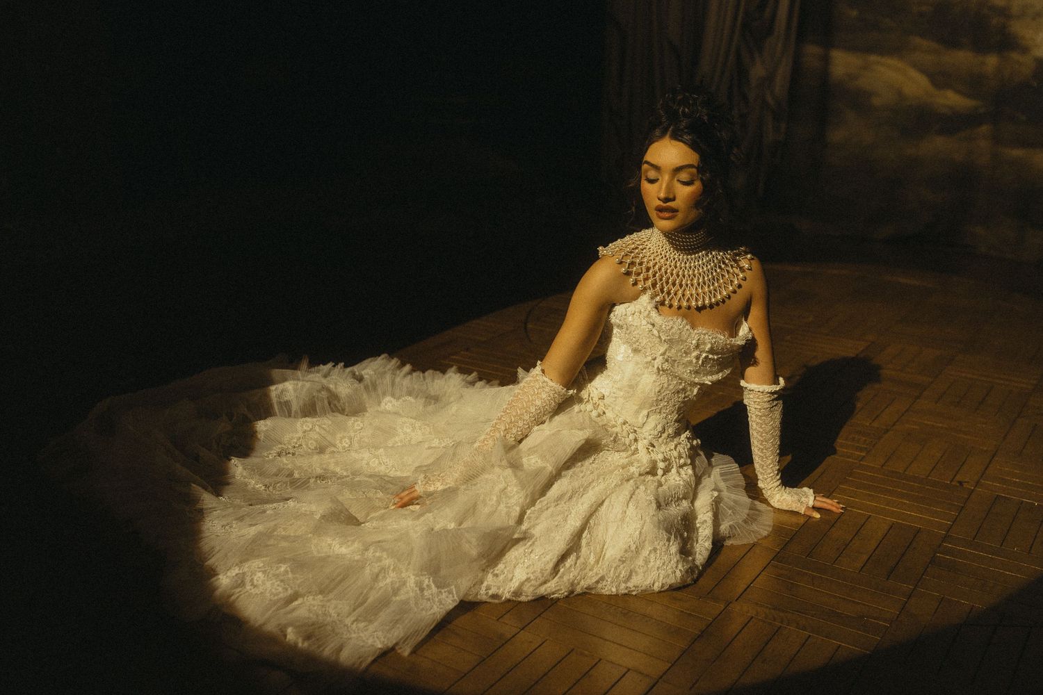 A bride in a white lace wedding gown poses gracefully on a hardwood floor in a dimly lit room.