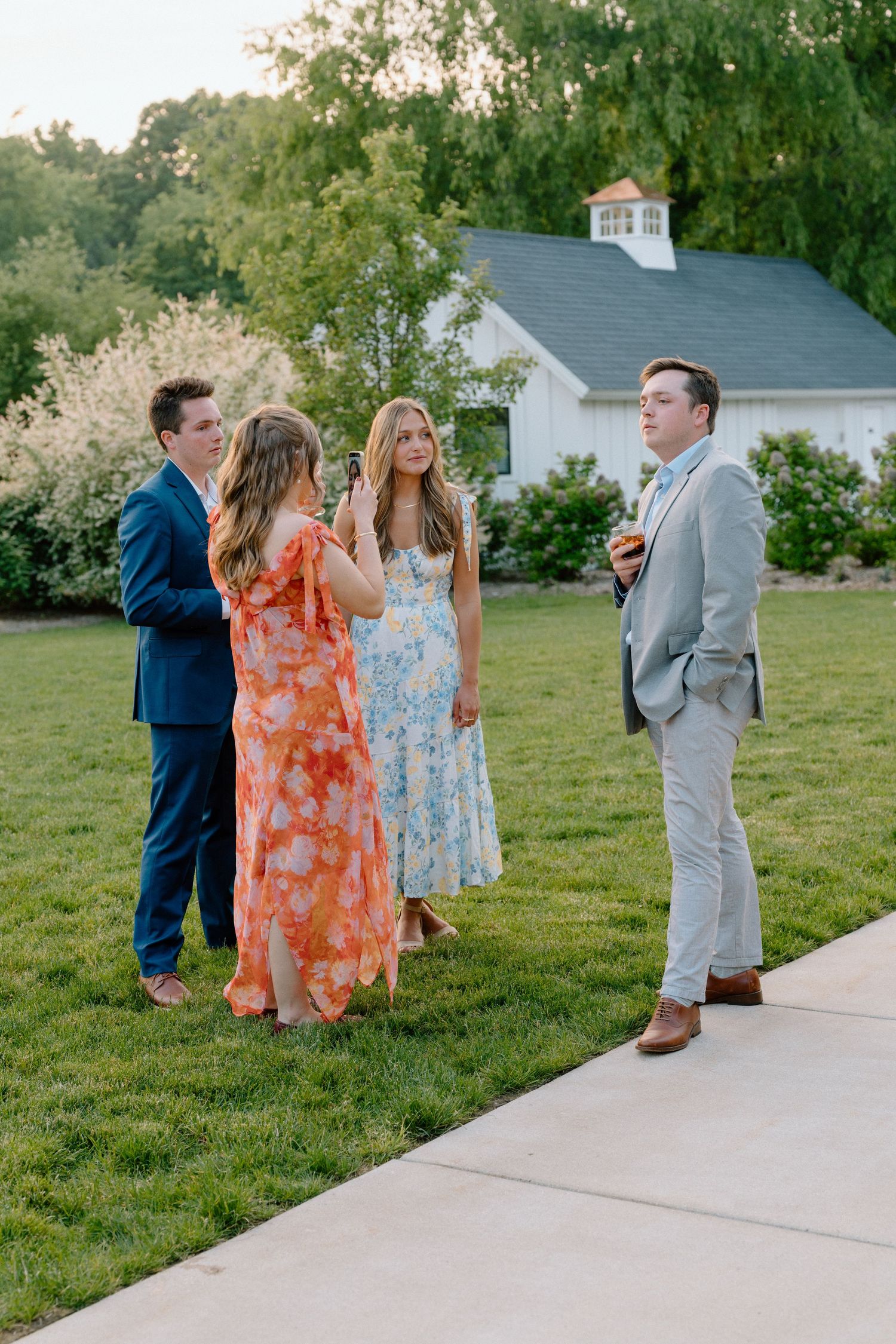 Wedding guests in colorful dresses gather on a lawn near white buildings.