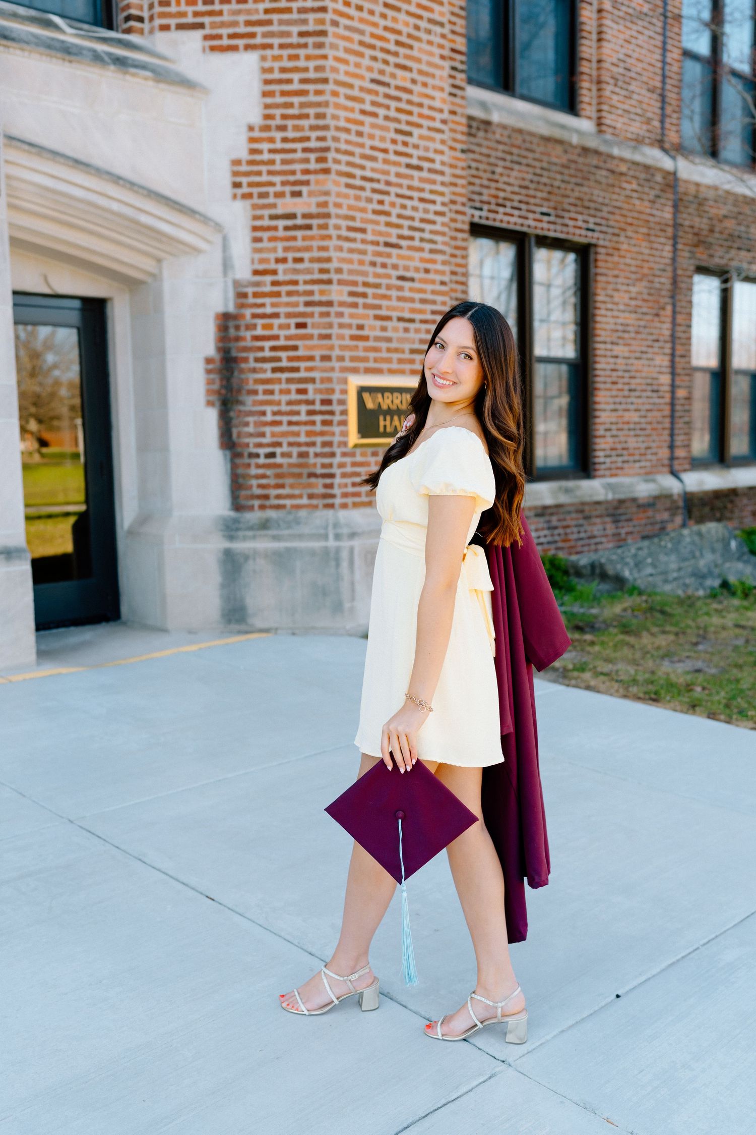 Young woman in white dress holding maroon graduation cap and gown outside brick university building.
