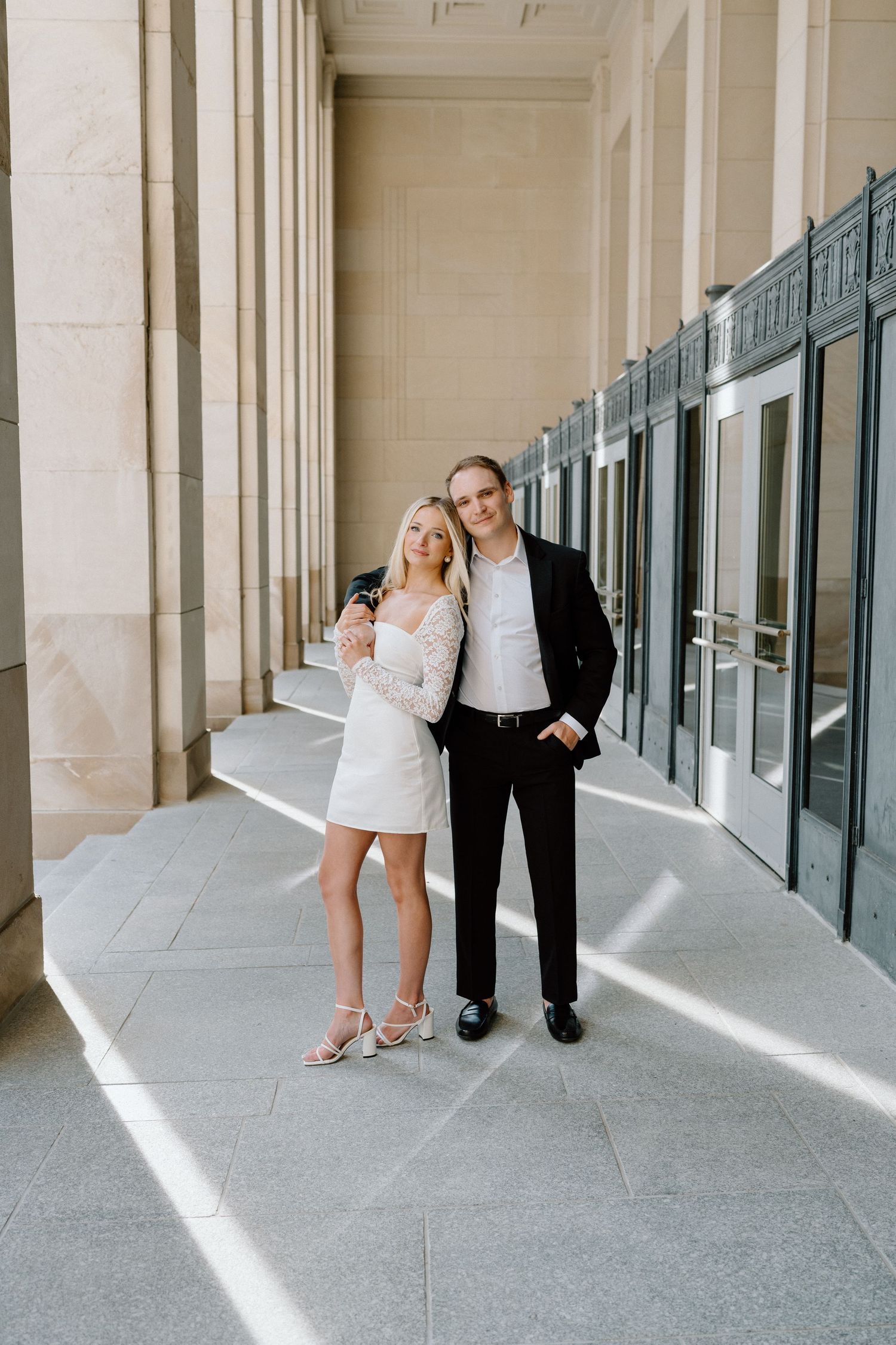 A couple poses together in an elegant colonnade with white columns and dramatic lighting casting shadows on the floor.