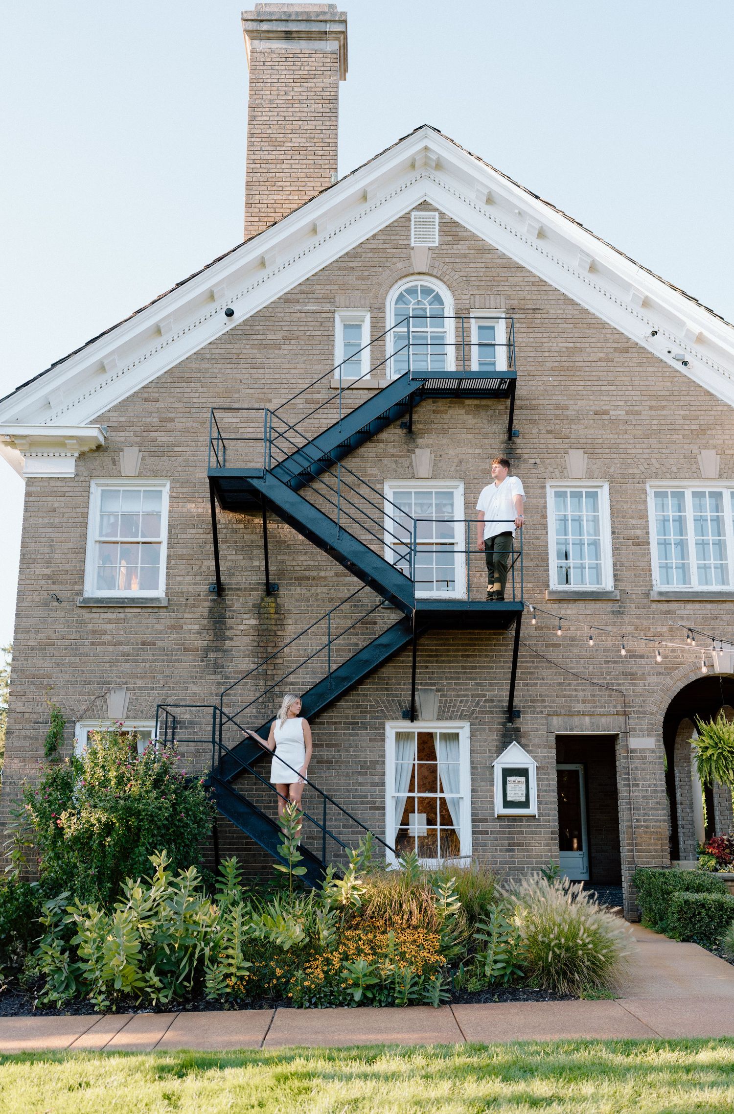 A couple poses on exterior metal stairs of a historic brick building.
