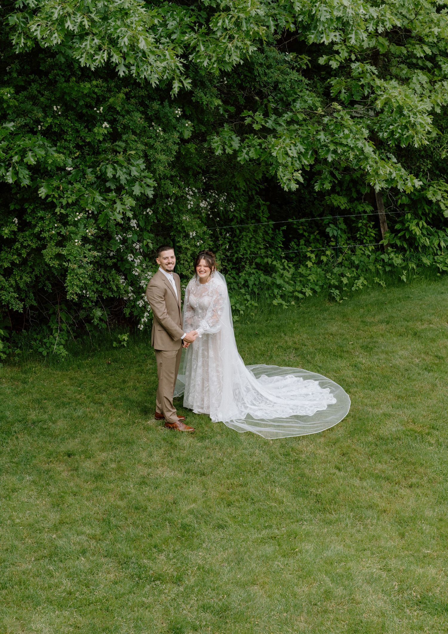 A couple in wedding attire shares a tender moment on a grassy lawn surrounded by lush greenery.