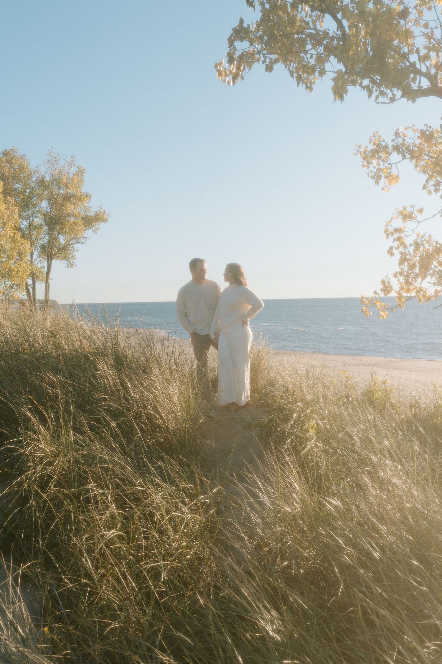 Two figures stand together on a grassy hillside overlooking the ocean at sunset.