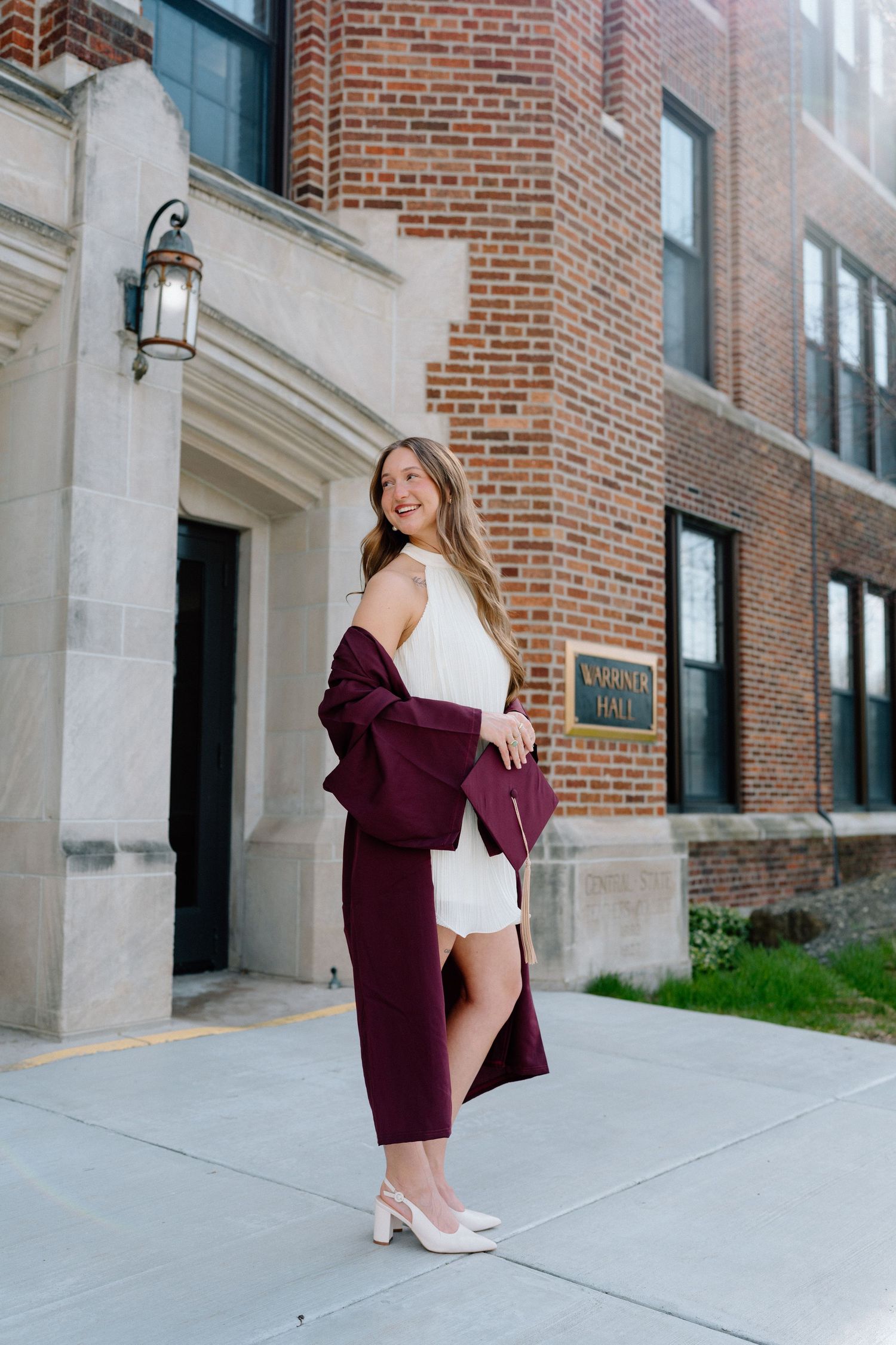 Young woman in white dress and burgundy blazer holding clutch, posing outside brick university building.