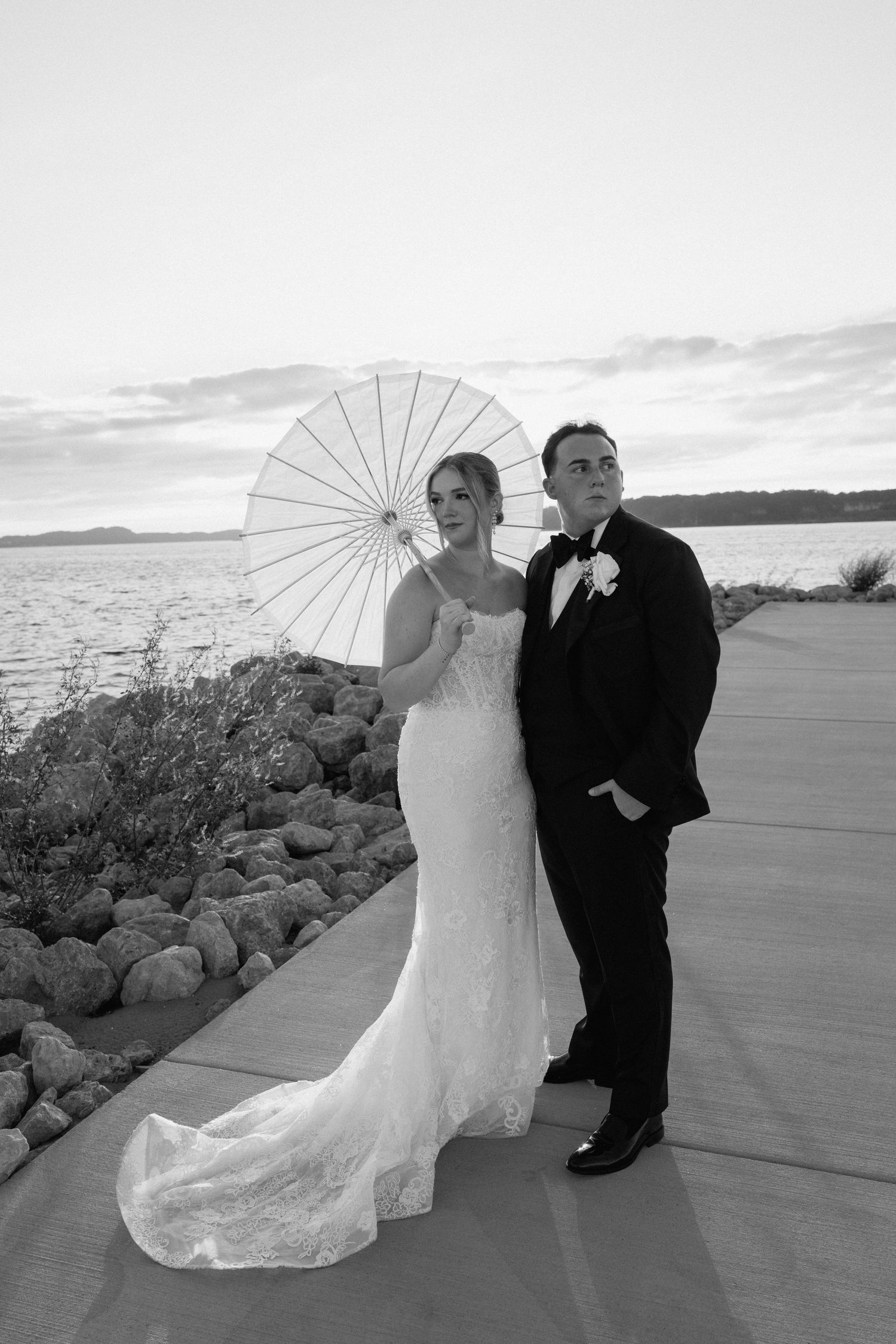 Wedding couple poses with white parasol umbrella along waterfront promenade in black and white photography series.