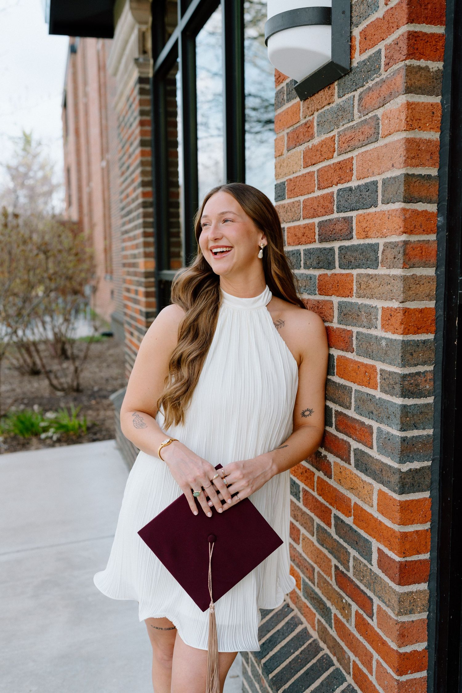 Smiling young woman in white halter dress holding a burgundy graduation cap, leaning against a brick wall.