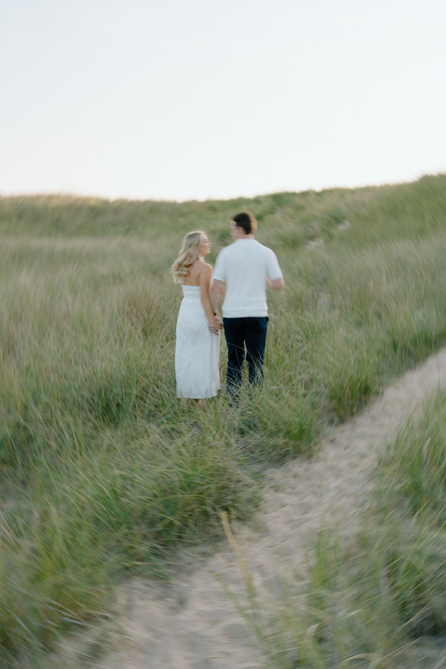 A couple walks hand in hand along a winding dirt path through tall grass on a hillside.
