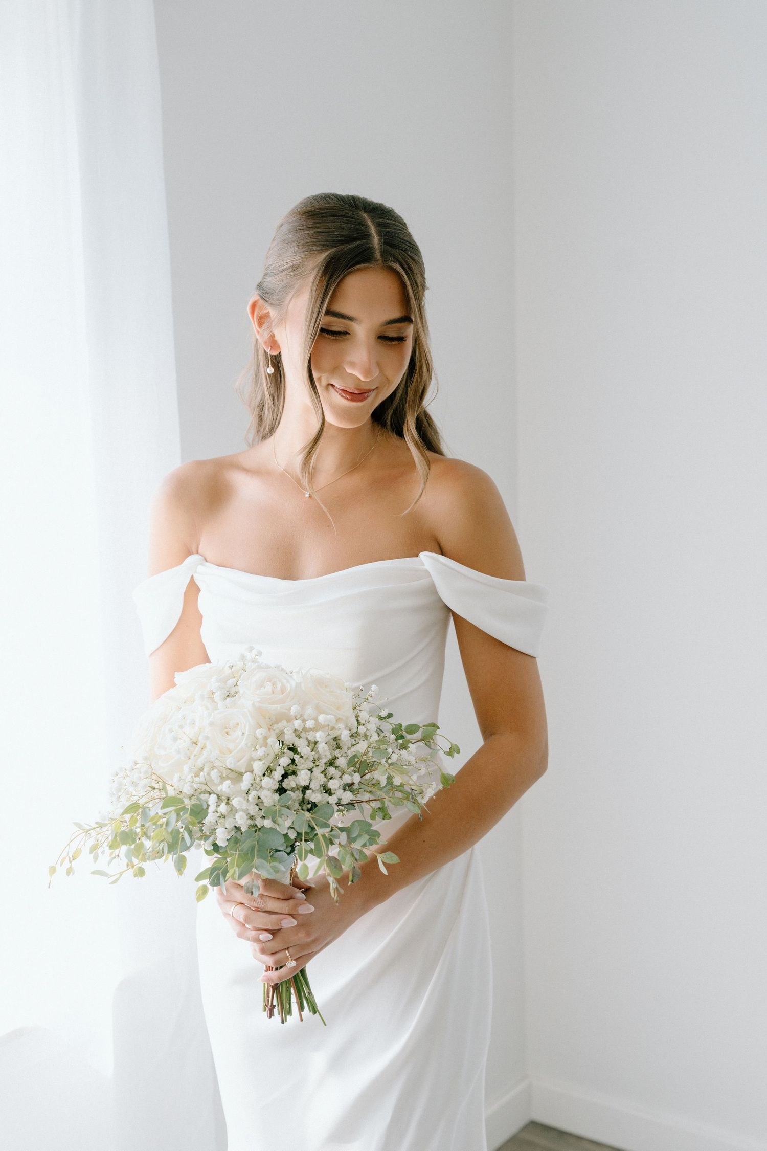 Bride in an elegant white off-shoulder gown holds a delicate bouquet of white flowers.