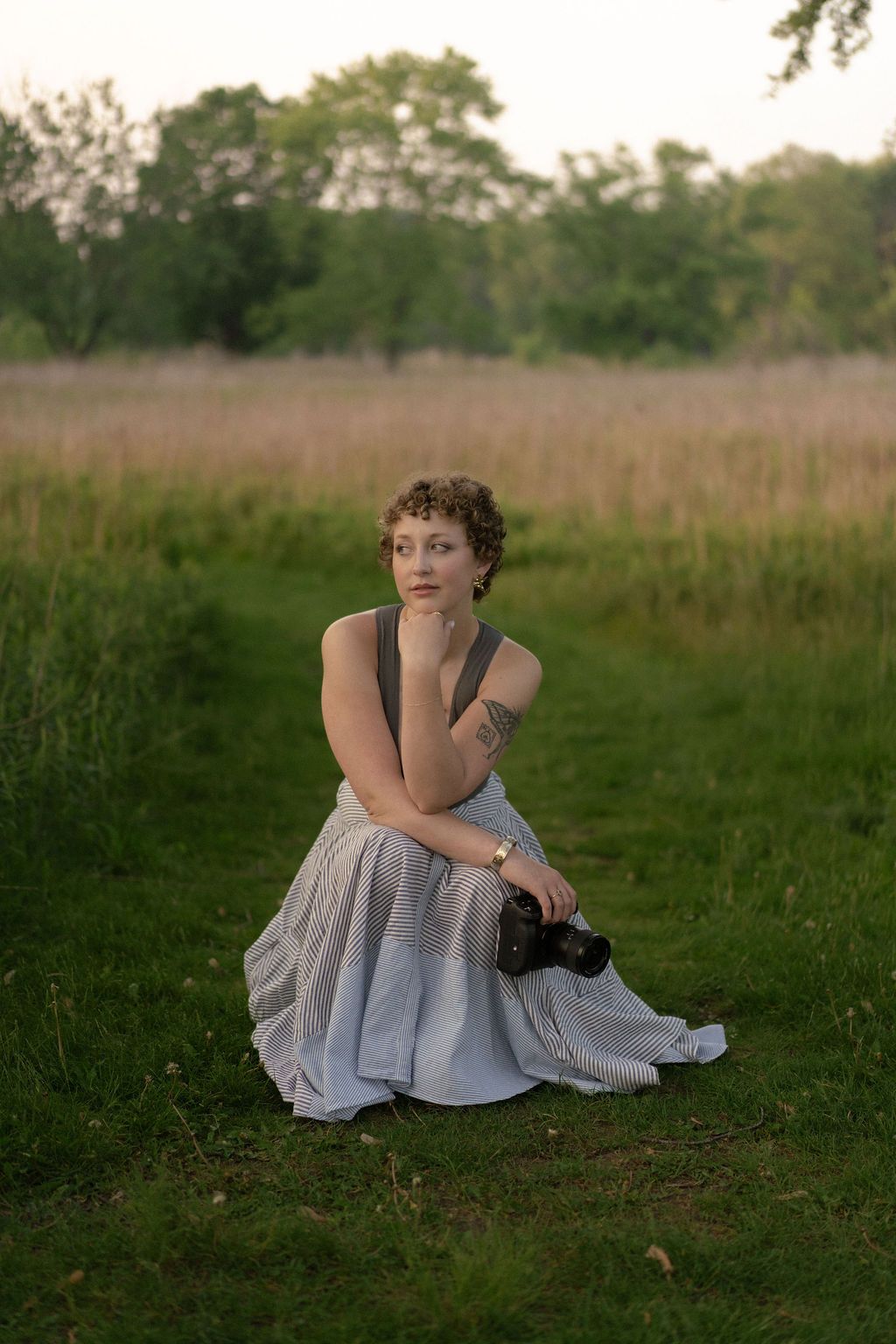 A figure in a light dress sits thoughtfully on grassy ground at dusk.
