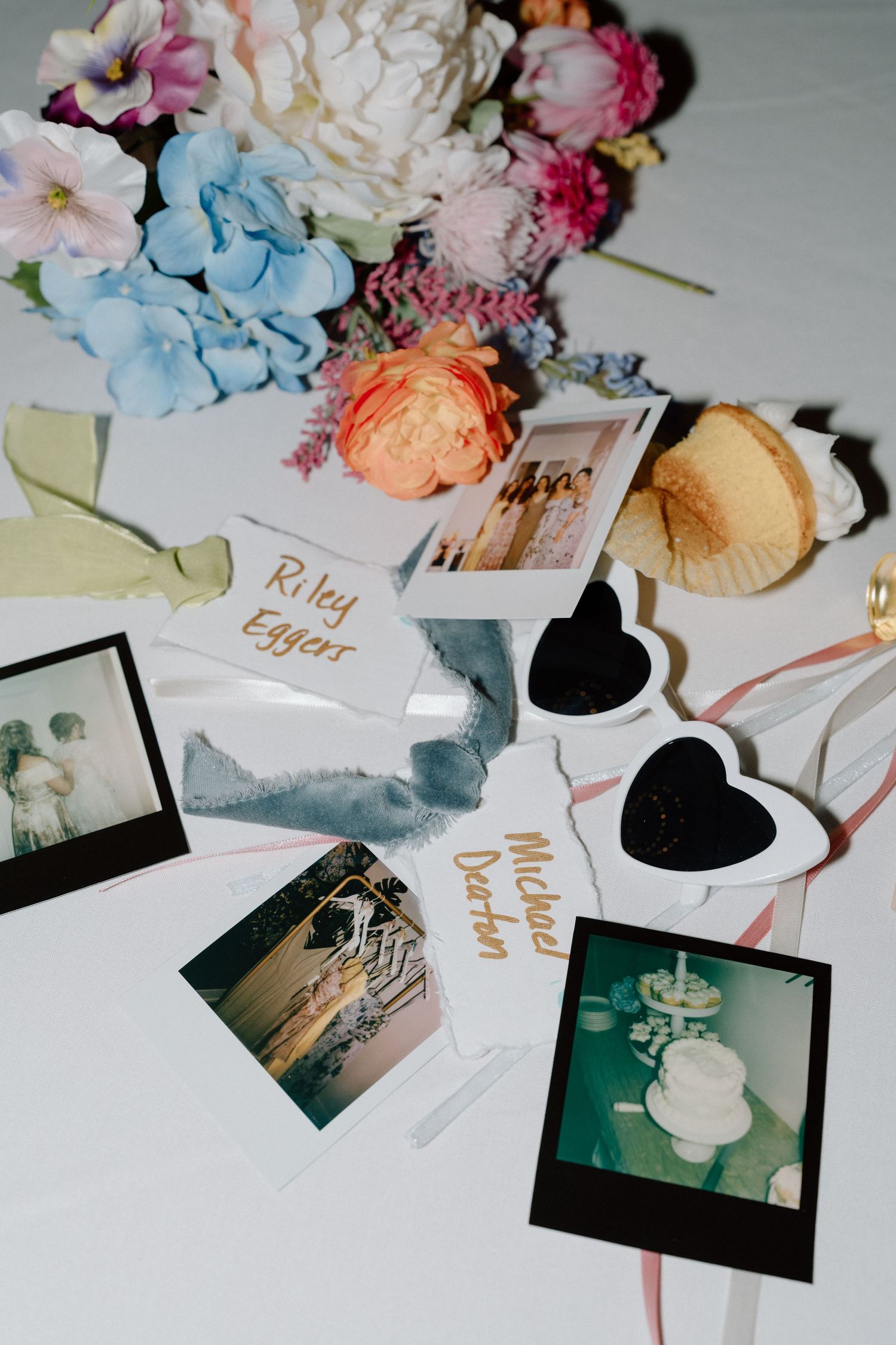 Wedding flatlay with colorful flowers, polaroid photos, and decorative elements on white surface.