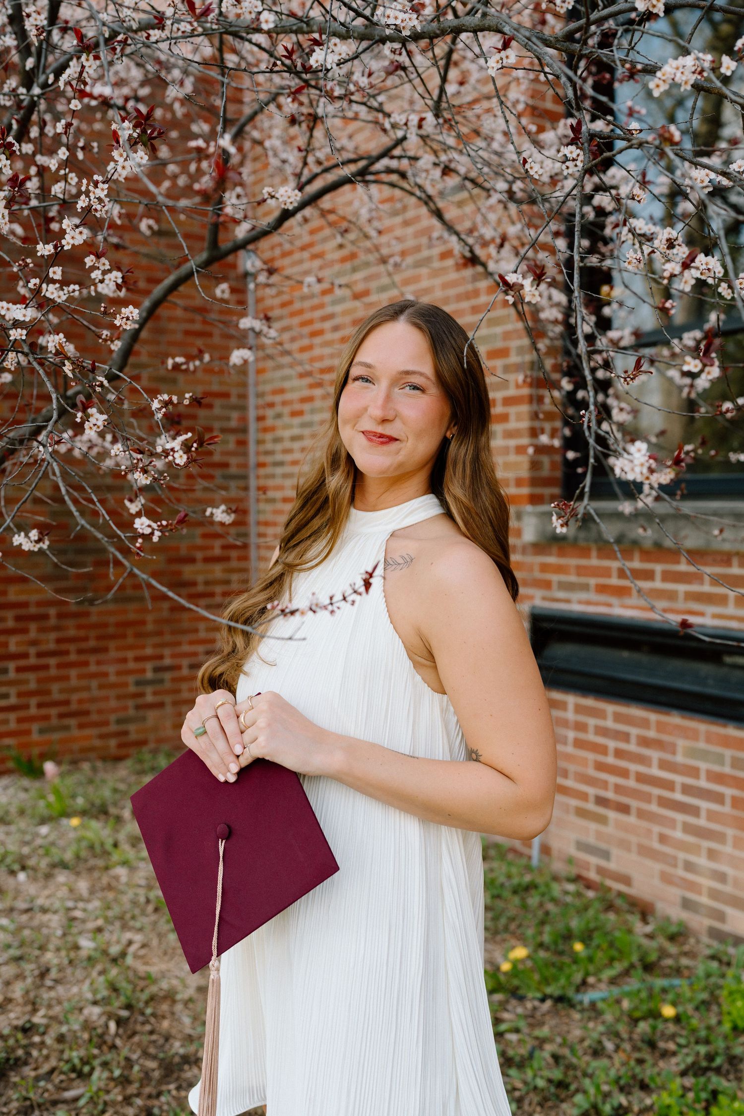 Young woman in white dress holding maroon graduation cap poses by cherry blossom tree near brick building.