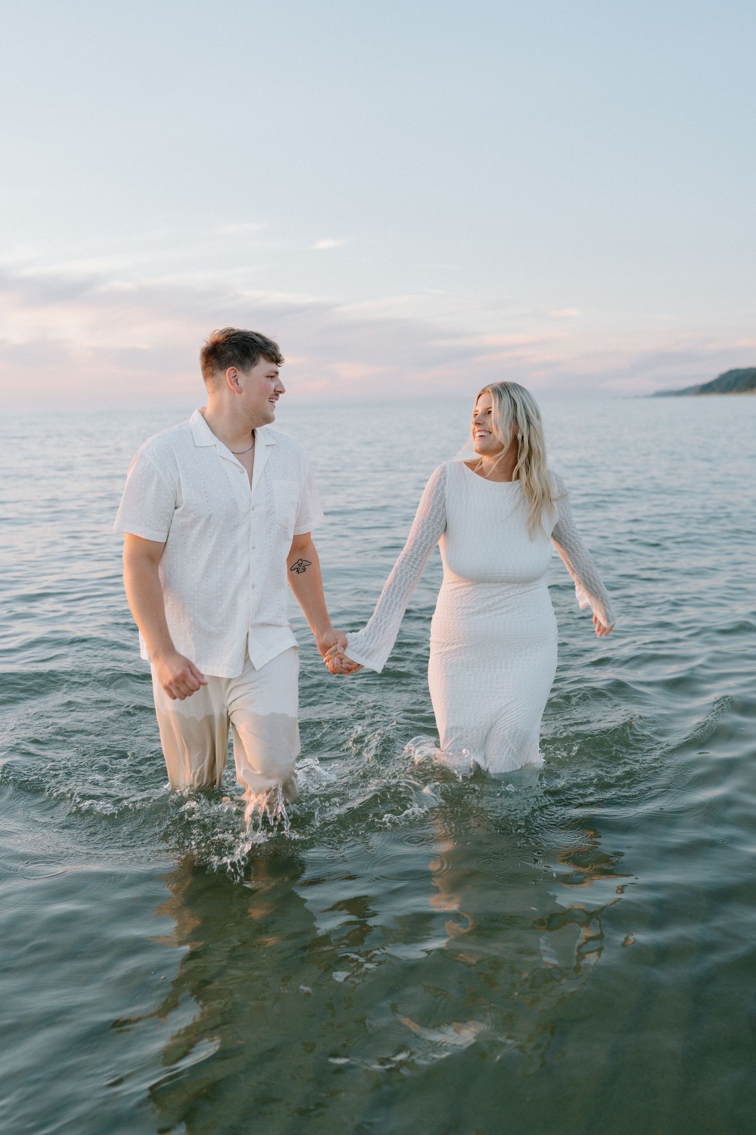 A couple splashes in shallow ocean water during sunset wearing white clothing.