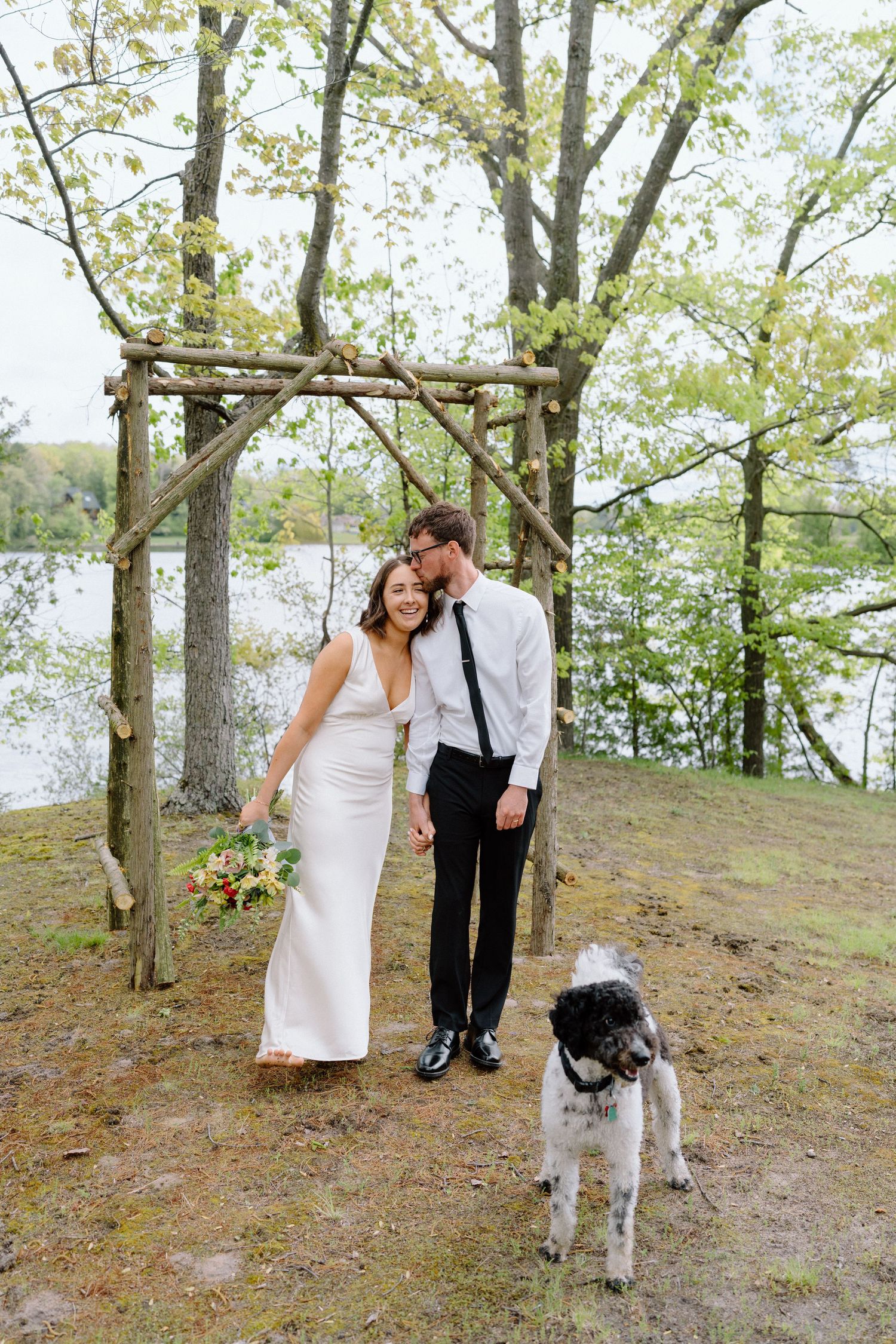 A couple and their dog stand together on a wooded hillside.