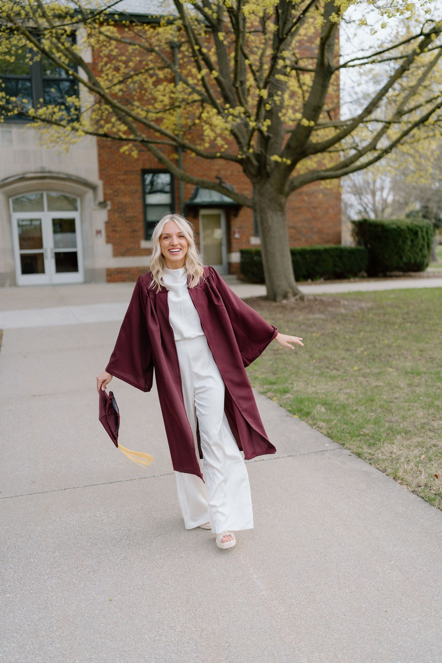 Graduate walking on campus pathway in burgundy graduation gown and white outfit.
