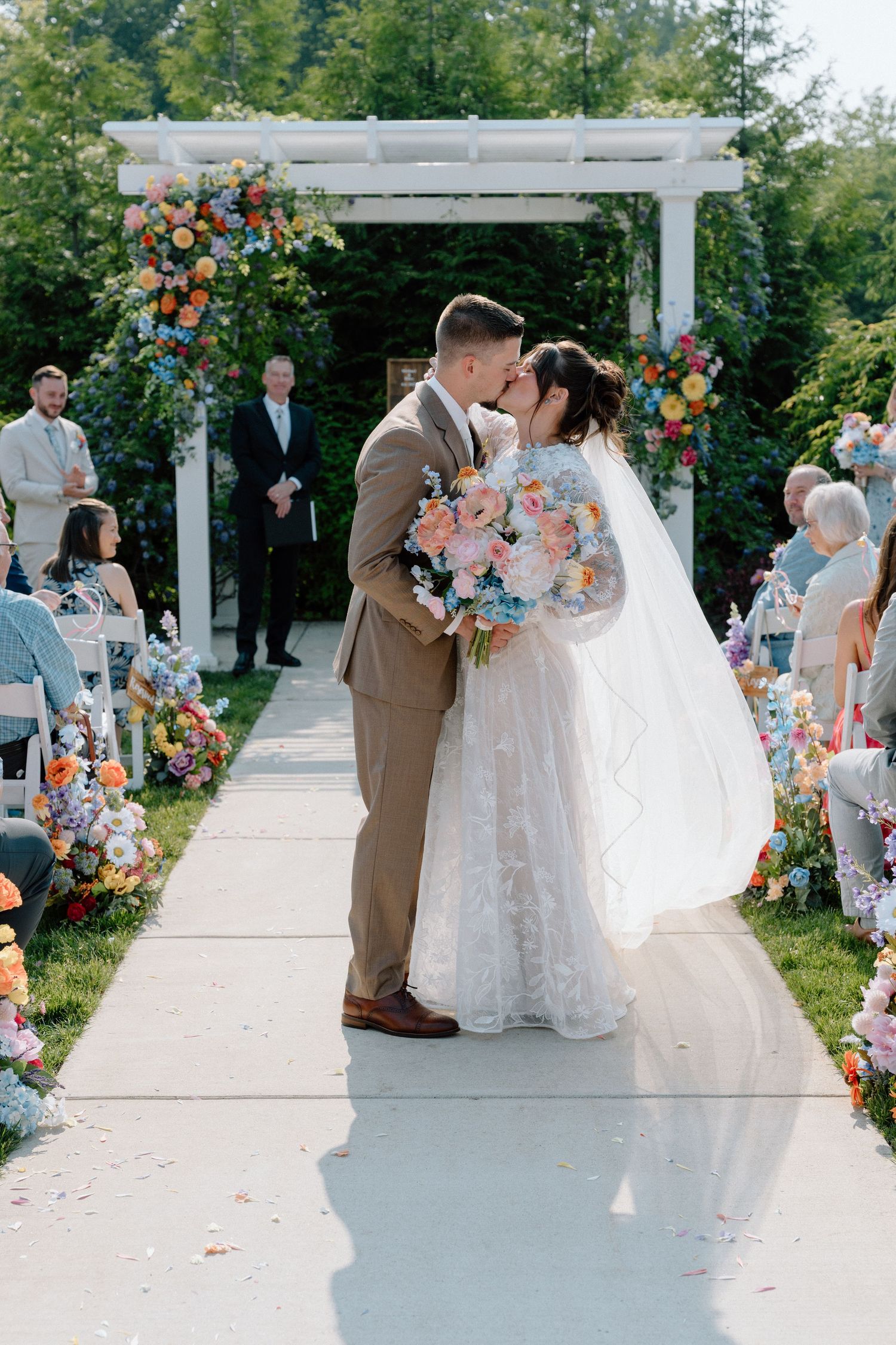 Wedding ceremony takes place under a white pergola decorated with vibrant orange and blue floral arrangements.