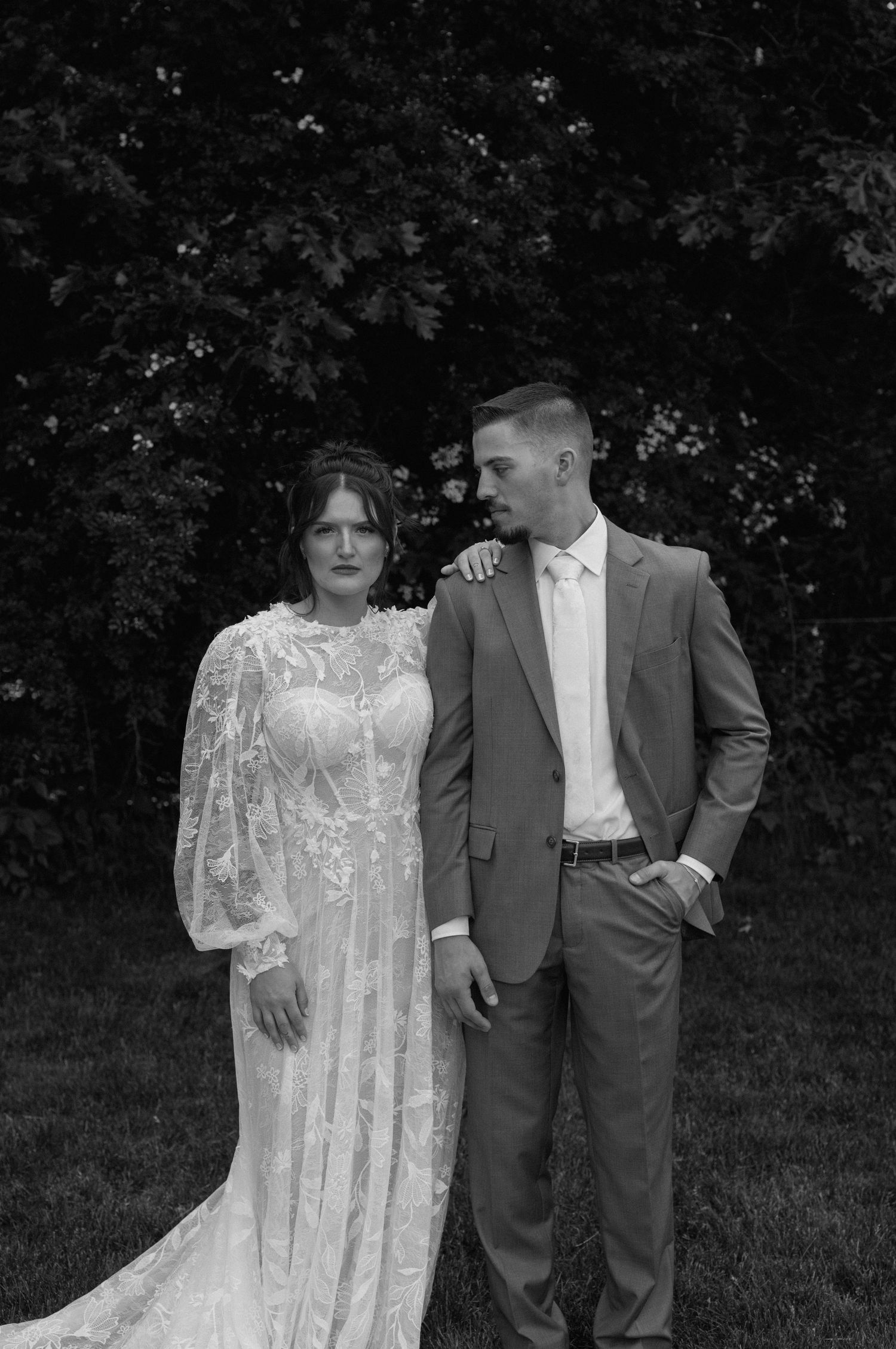 Black and white portrait of couple in wedding attire standing against natural backdrop.
