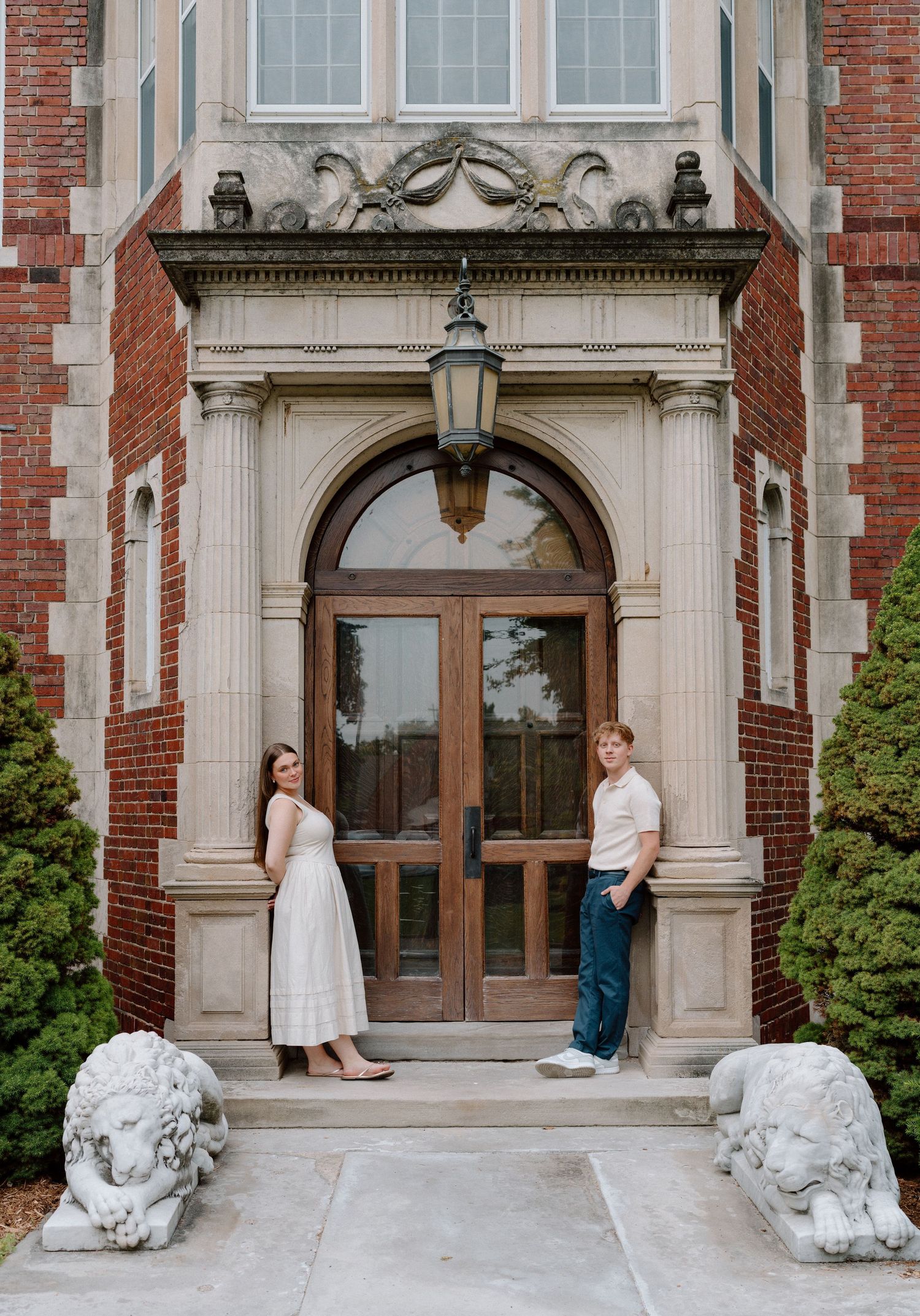 Two people posing at ornate building entrance with stone lions and brick facade.