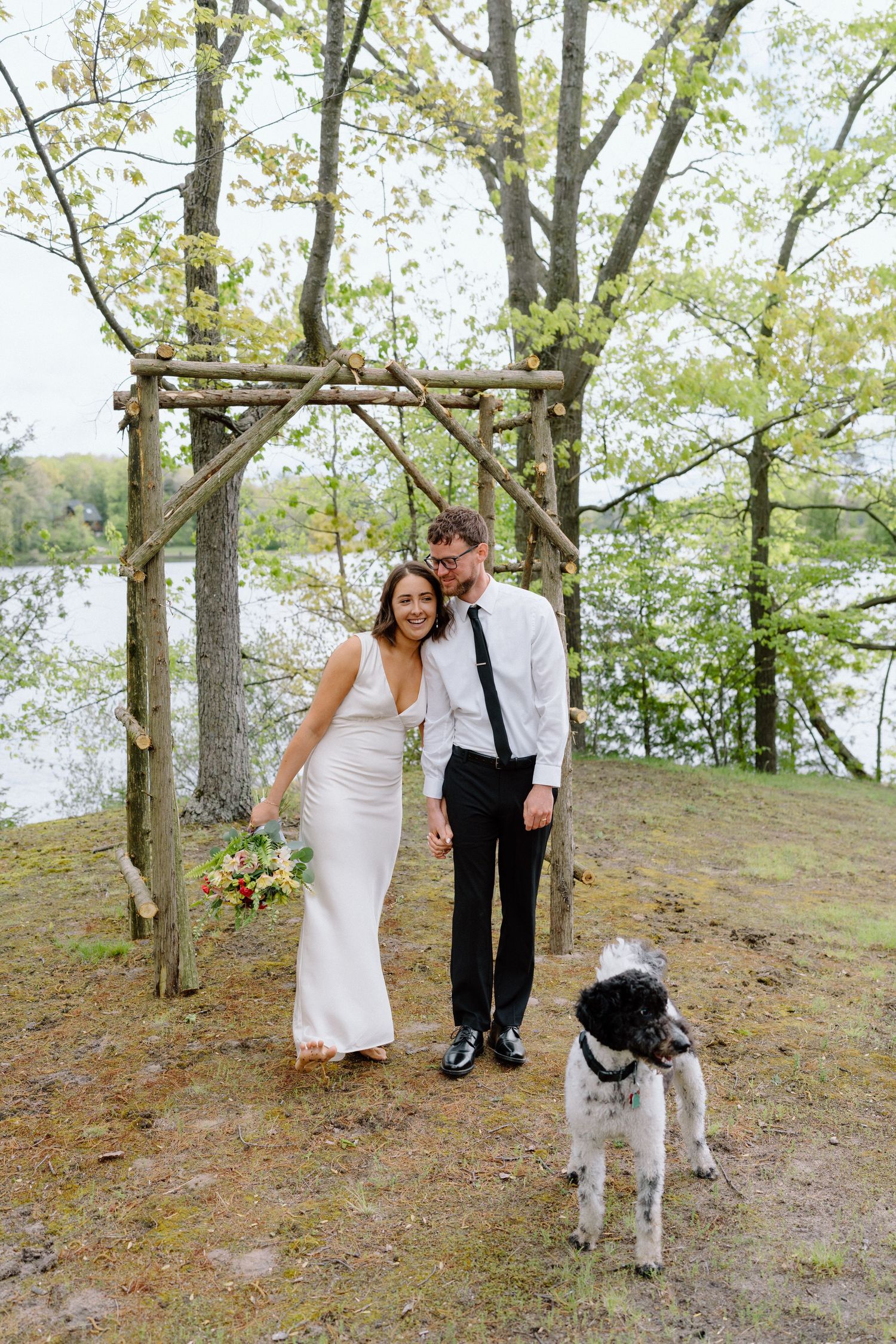 A couple poses with their dog on a hill with trees in the background.